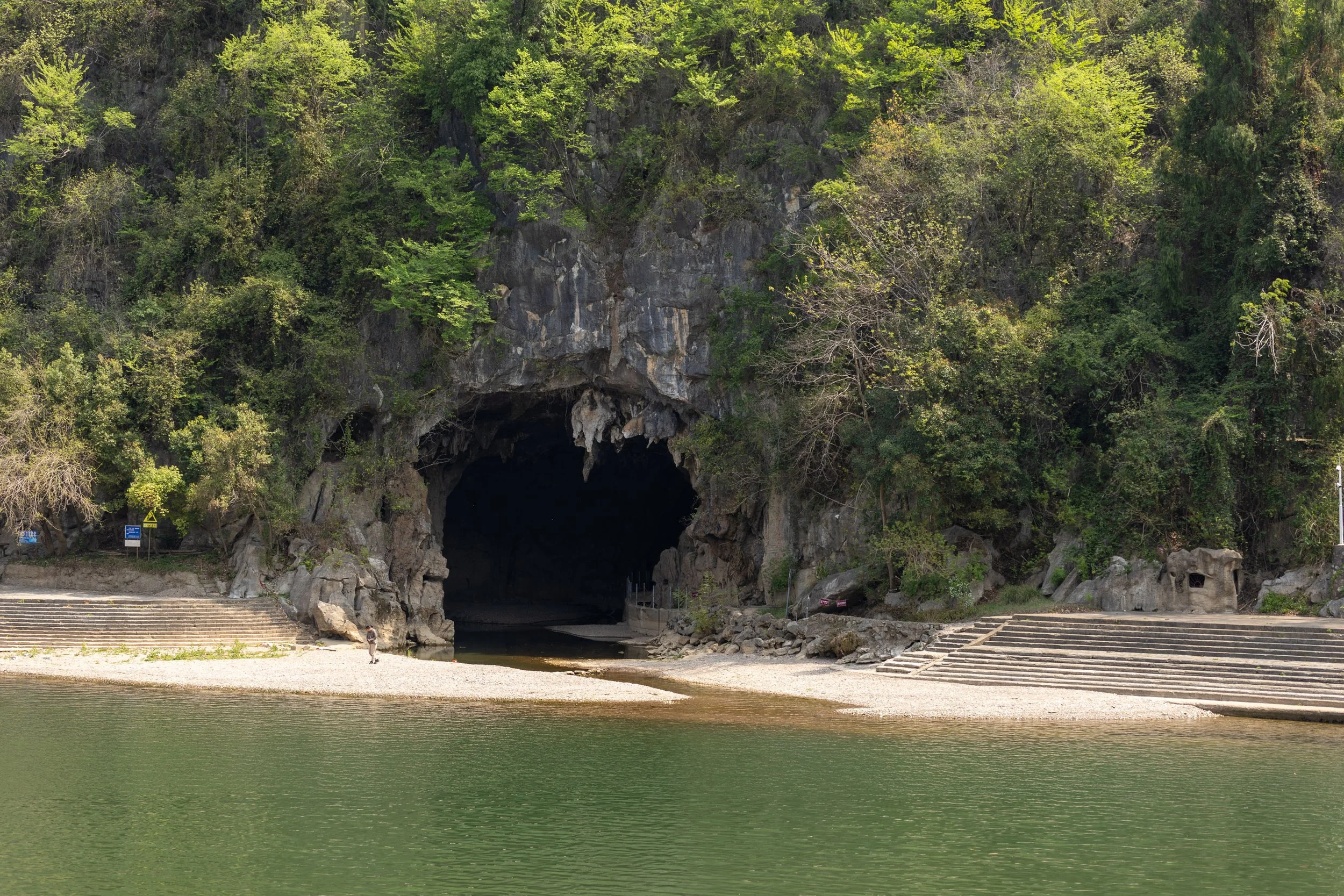Cave entrance surrounded by lush greenery, with stone steps and a small beach leading to a body of water.