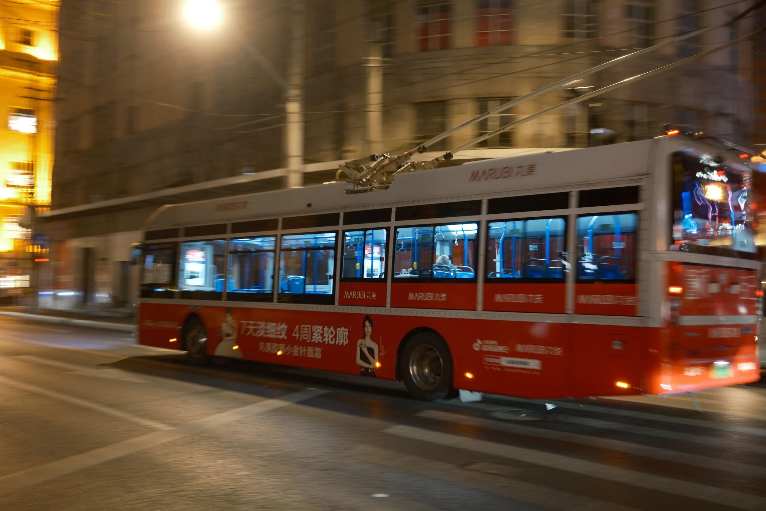 Red trolleybus moving on a city street at night, with advertisements on the side and headlights illuminating the road.