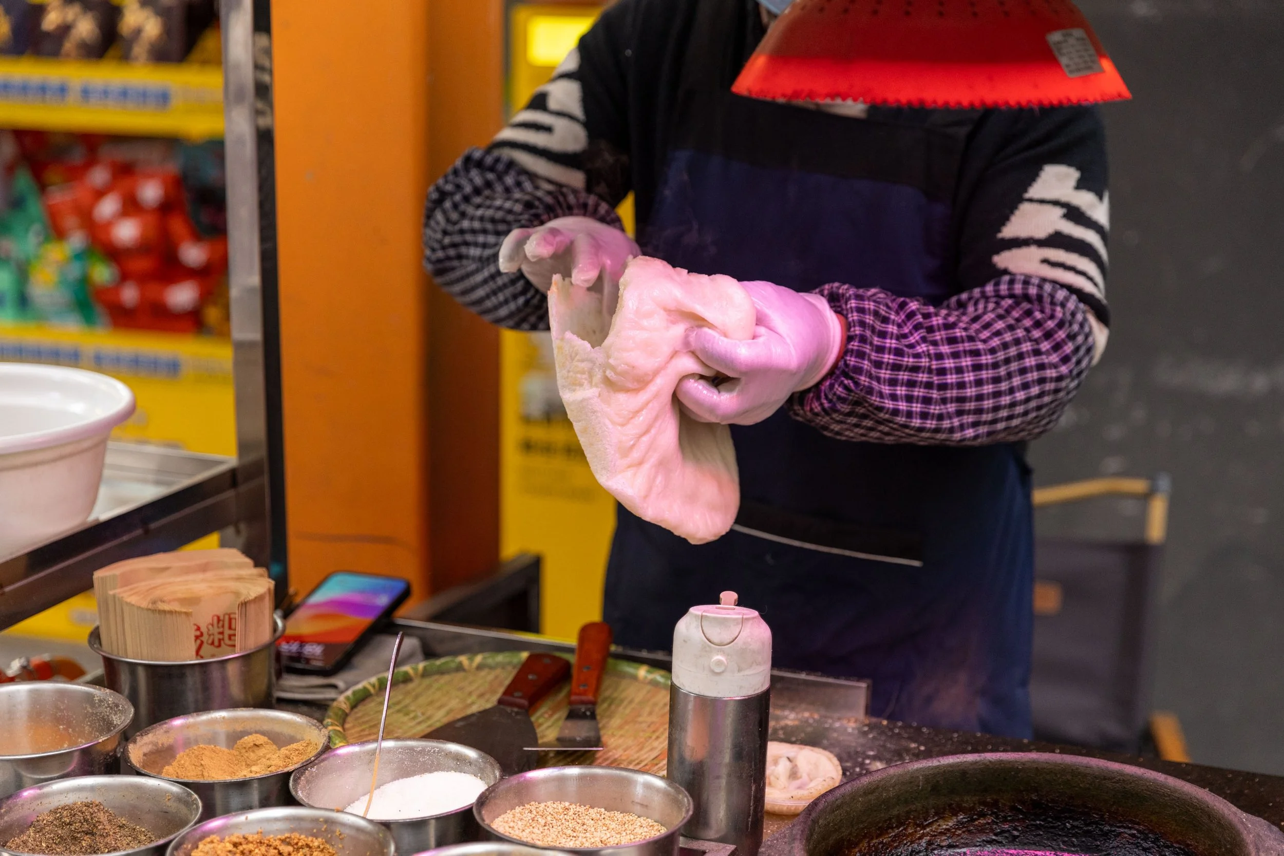 Person wearing gloves handling dough at a food stall with various seasonings in bowls.