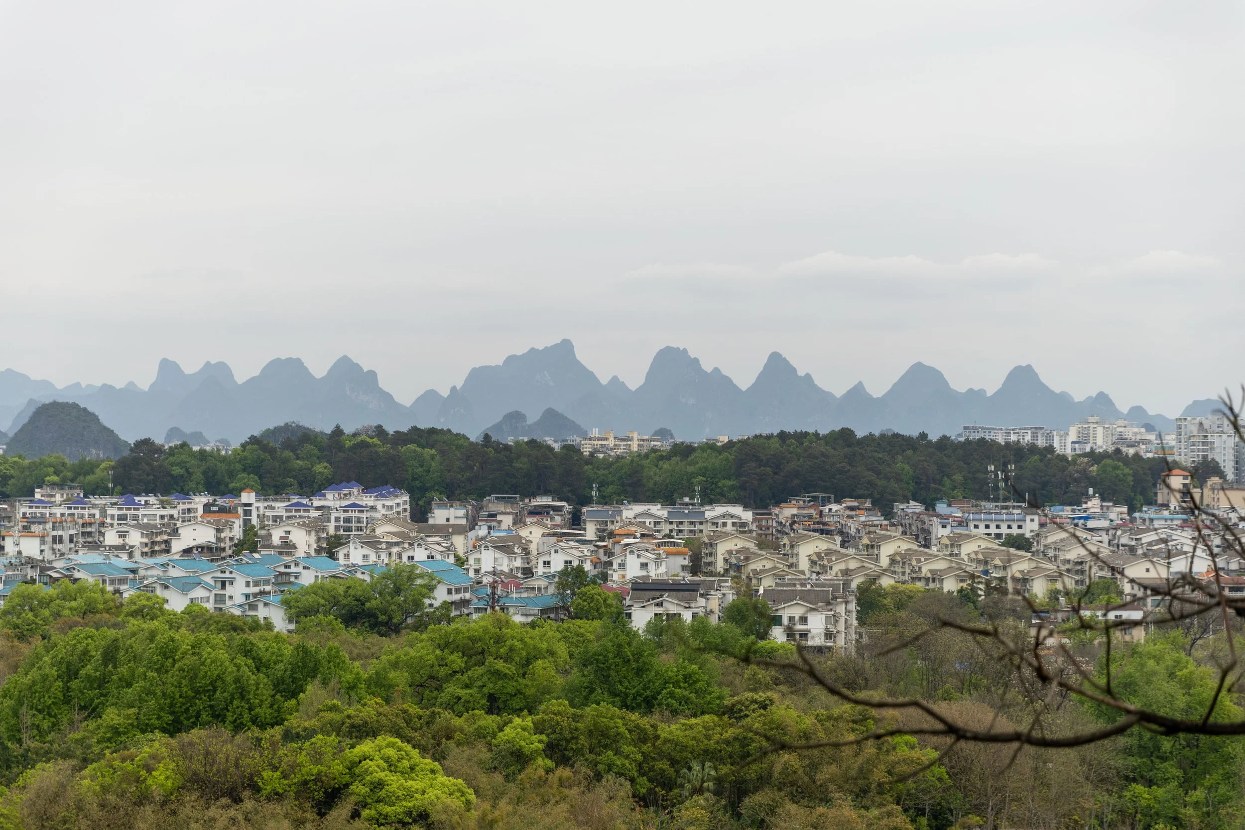 Cityscape with buildings, greenery, and mountainous backdrop under cloudy sky.