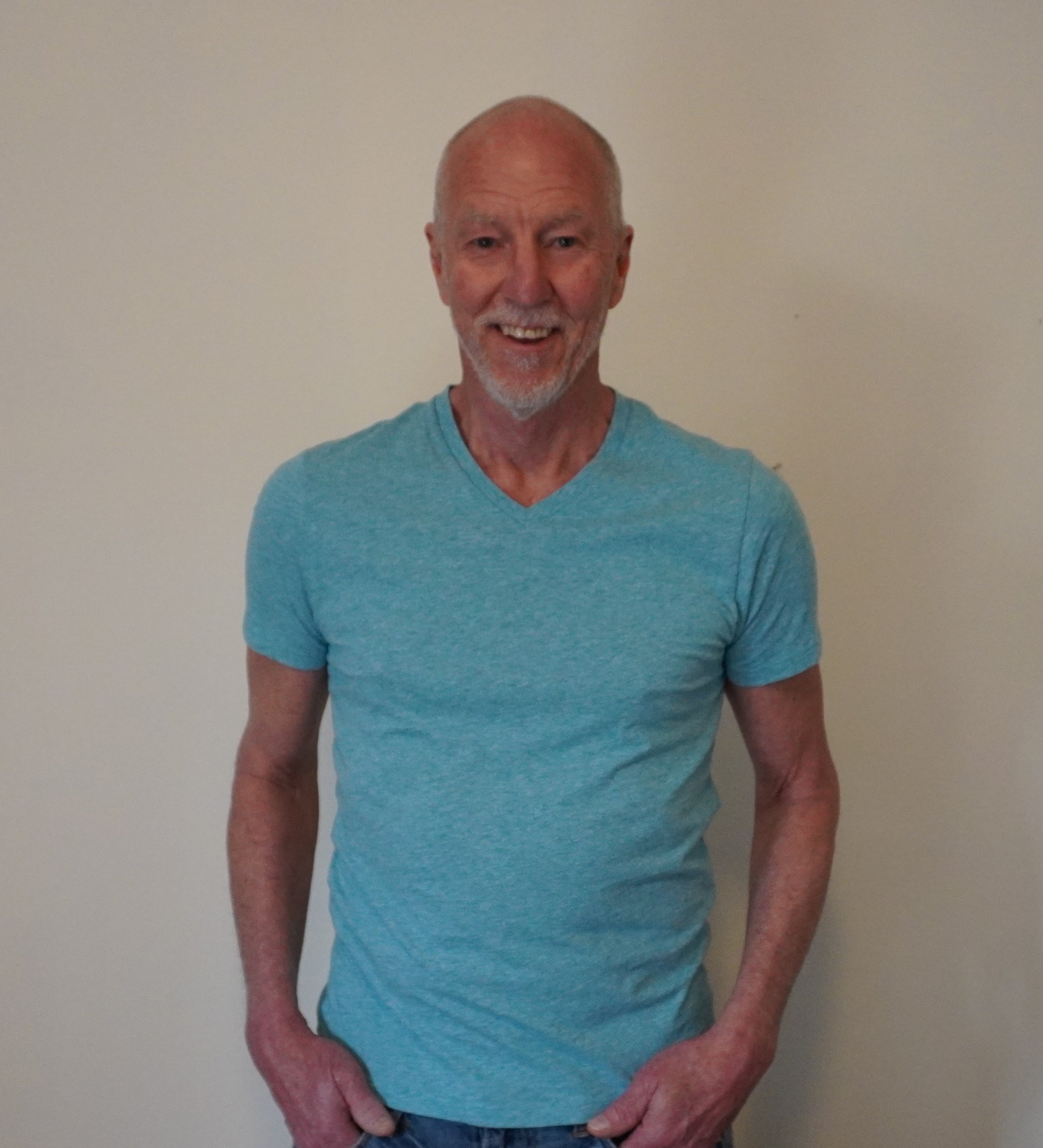 An older man with a bald head and gray beard smiling, wearing a light blue V-neck T-shirt, posing against a plain off-white wall.