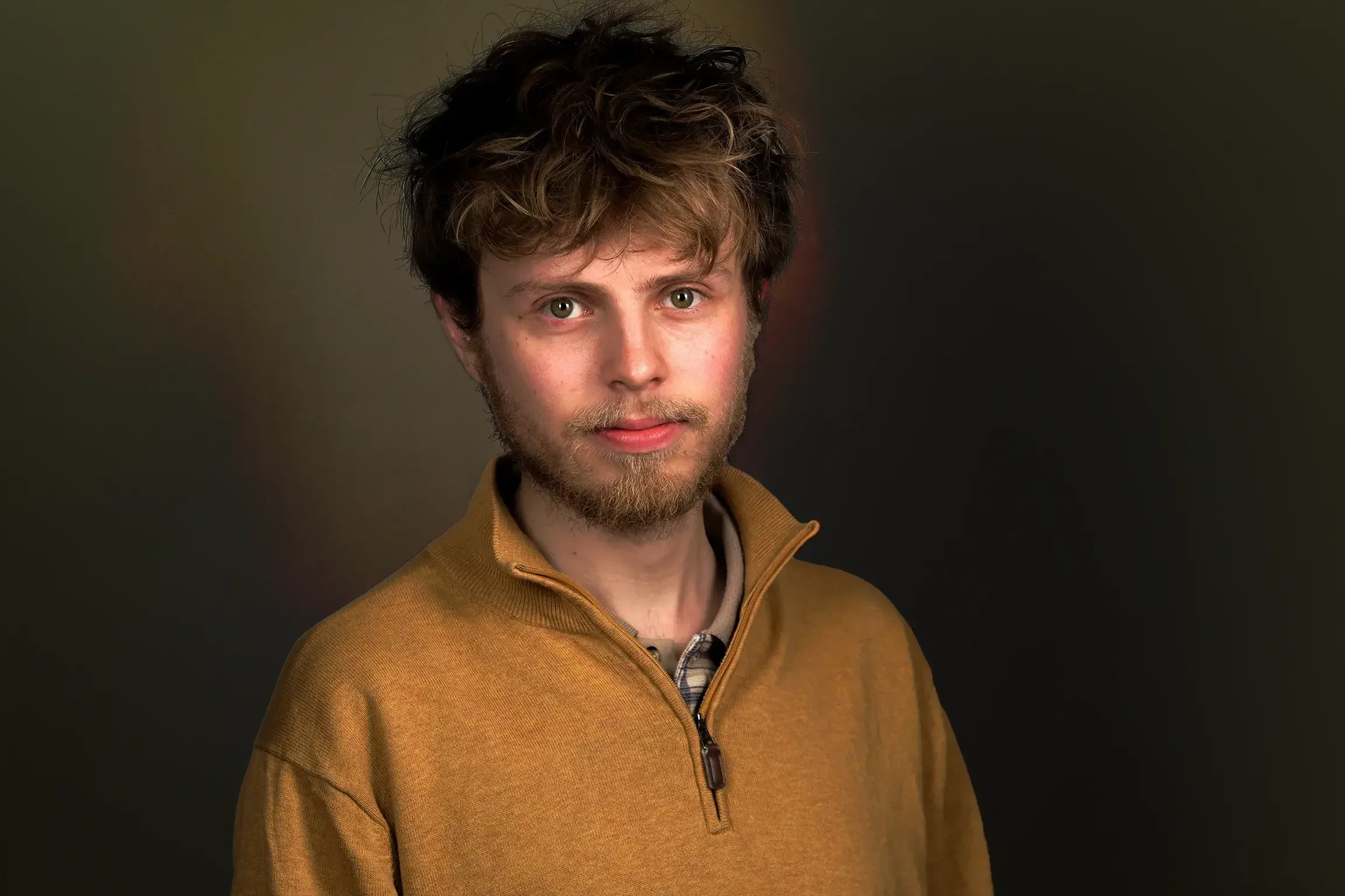 Portret van een jonge man (Daniël) met groene ogen, een lichte baard en warrig, donkerbruin haar, die een bruinachtige gele sweater draagt tegen een donkere achtergrond.