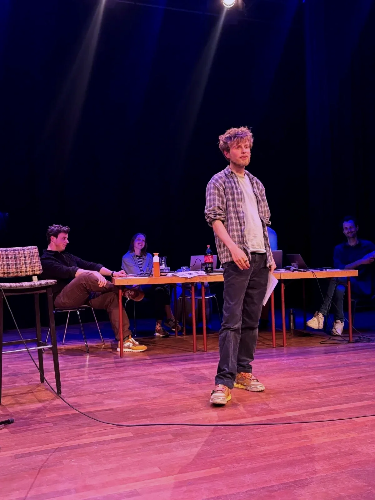 Man (Daniël) staat op een podium tijdens een groeps-optreden, terwijl drie mensen achter een tafel zitten, lachend en kijkend naar hem. Op tafel liggen diverse drankjes en laptops. Het decor is donker met gekleurde lichten.