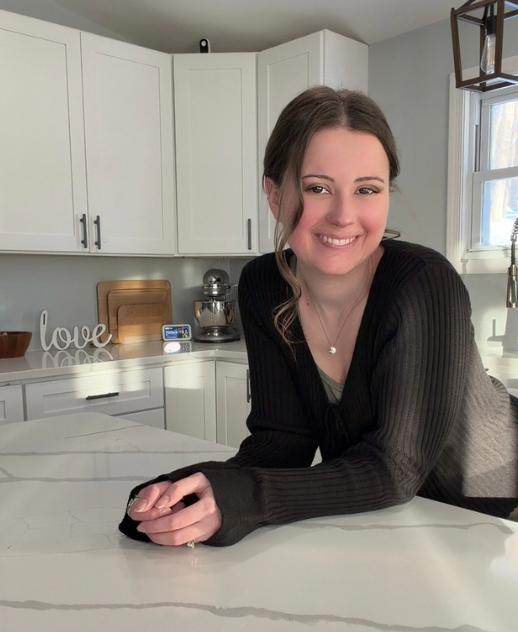 a friendly woman with brown hair in a black sweater leaning on a white kitchen counter