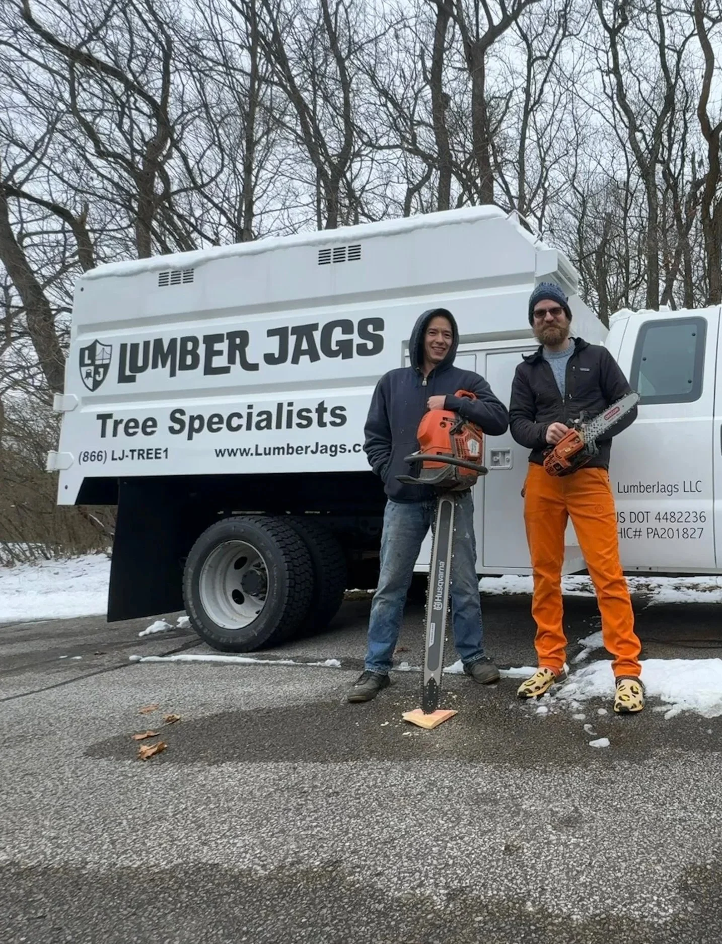 Two men standing in front of a lumber truck outdoors on a snowy day, holding chainsaws, smiling at the camera.