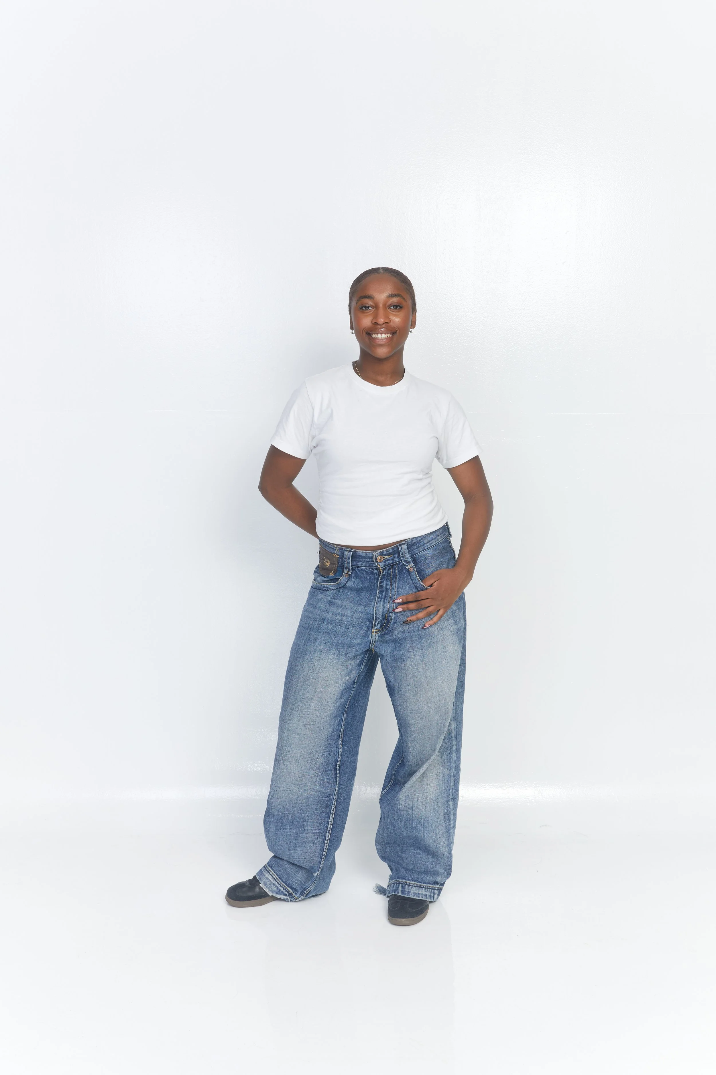 A young woman smiling, wearing a white t-shirt and baggy blue jeans, standing against a plain white background.