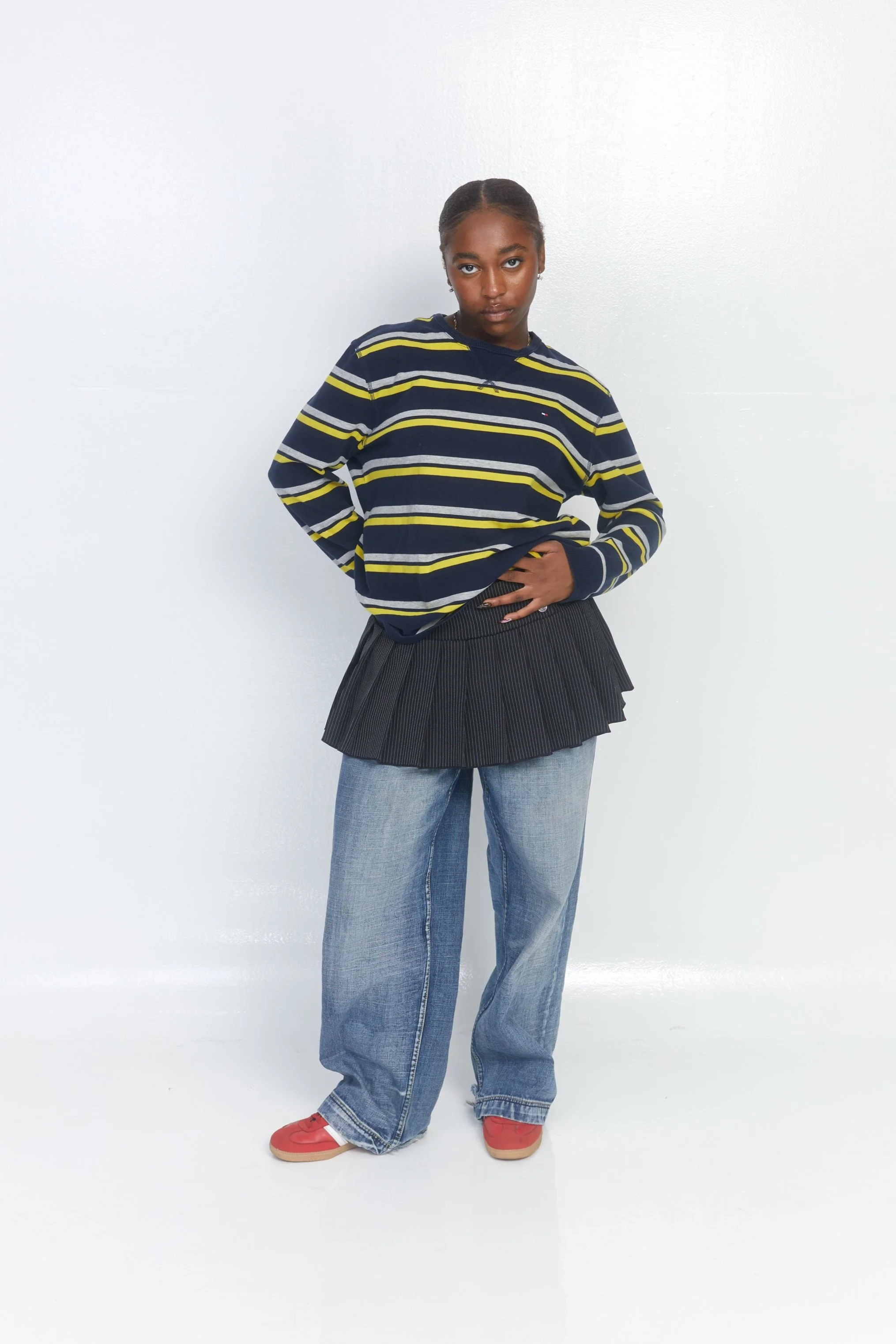 Young woman standing against a white wall, wearing a striped navy and yellow sweatshirt, pleated black skirt, loose blue jeans, and red shoes.