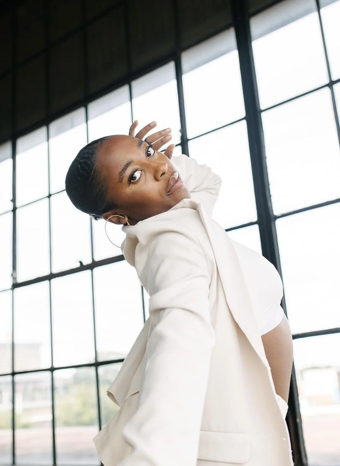 A woman with braided hair and hoop earrings wears a beige blazer over a white top, posing with her hand near her head in front of large industrial windows.