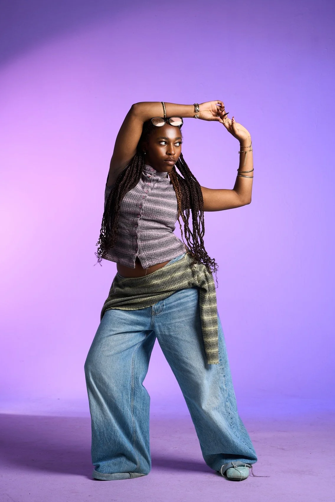 A young woman with long braids posing with her arm raised, wearing glasses on her head, a sleeveless top, wide-leg jeans, and a checkered shirt tied around her waist against a purple background.