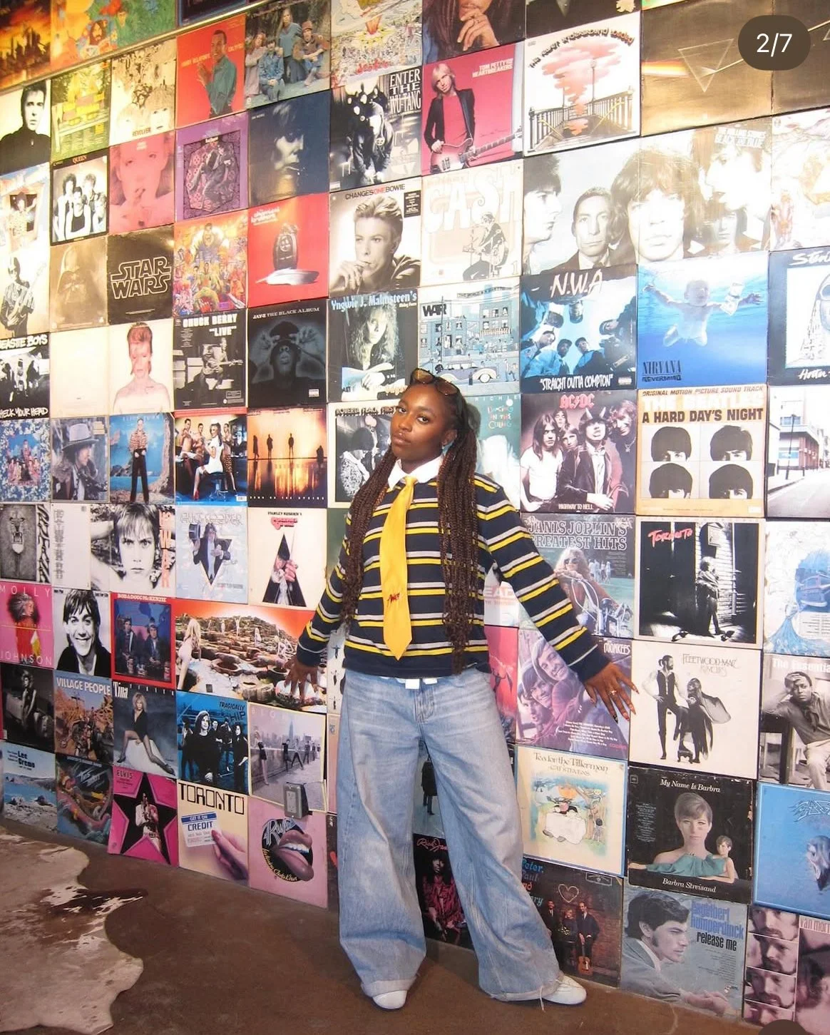A woman with  a striped navy blue and yellow sweater with a yellow tie poses with her arms outstretched in front of a wall decorated with album covers.