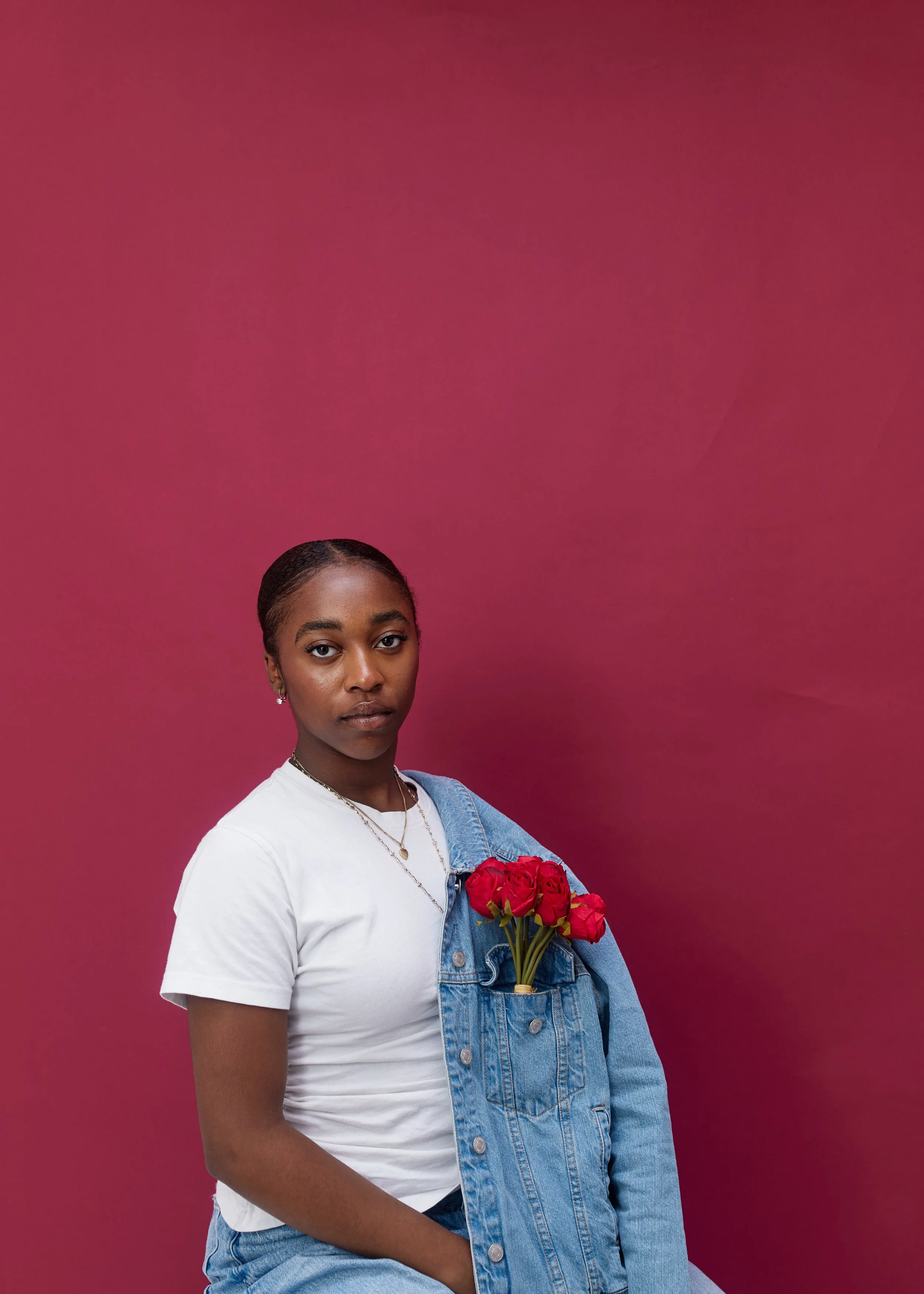 A young woman wearing a white t-shirt and denim jacket carrying a bouquet of red roses against a pink background.