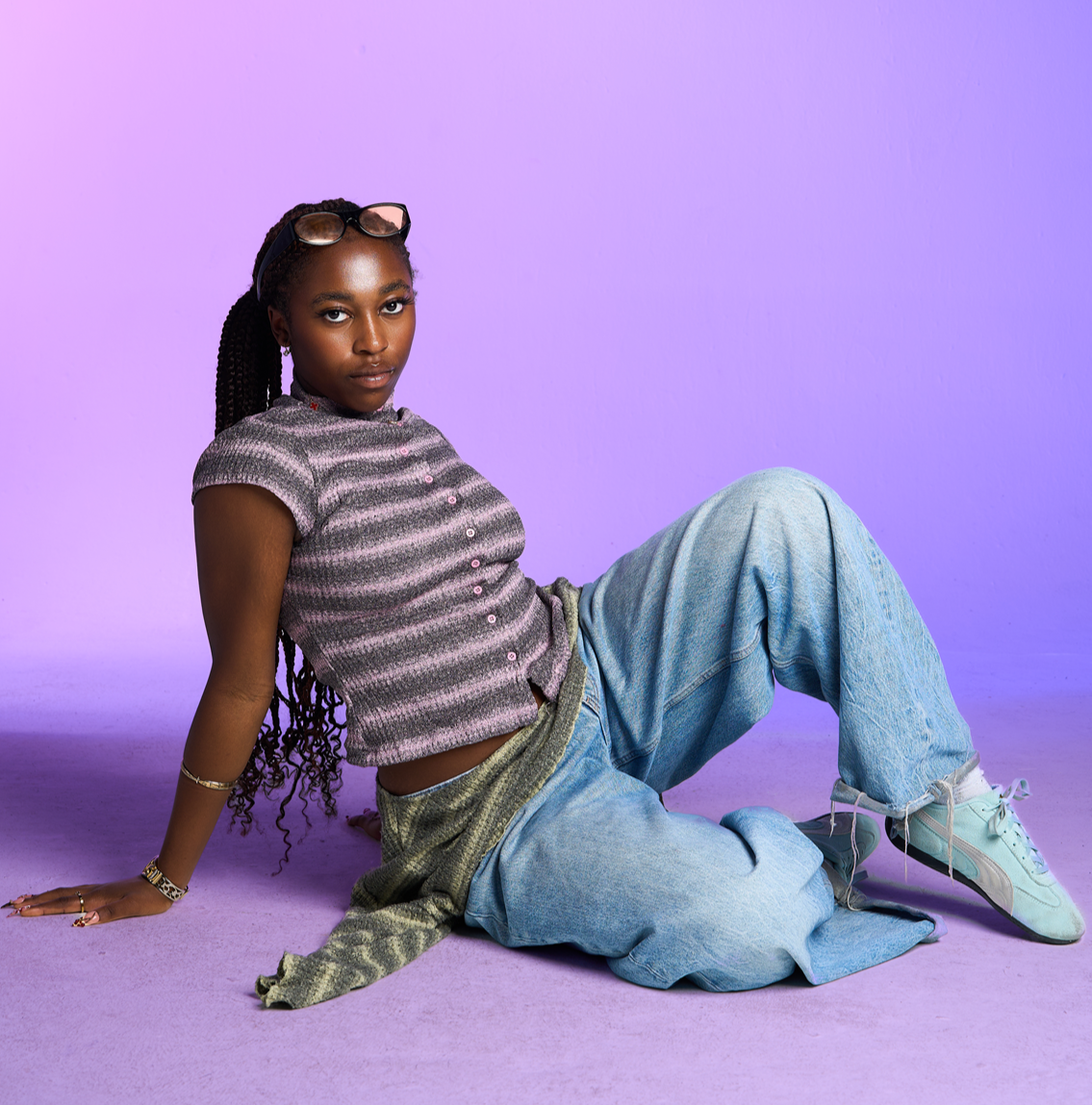 A young woman with braided hair and glasses on her head, sitting on a purple surface with a matching purple background. She is wearing a striped, short-sleeve button-up shirt, loose light blue jeans, and sneakers, and is looking directly at the camera.