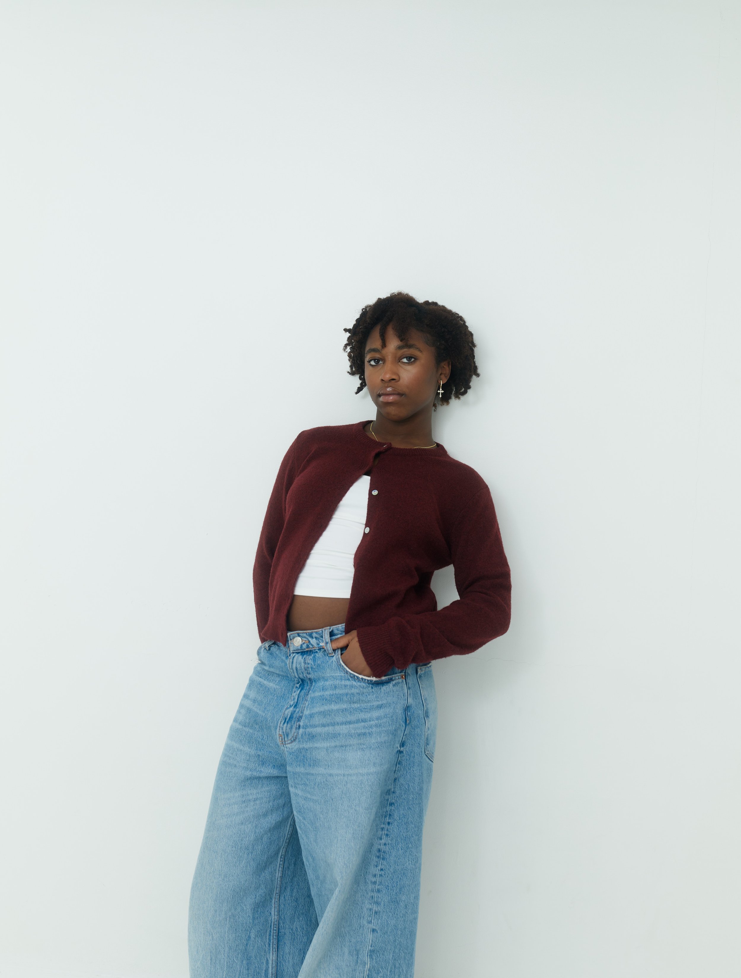 A young woman with curly hair leaning against a white wall, wearing a maroon cardigan, white crop top, and high-waisted blue jeans.
