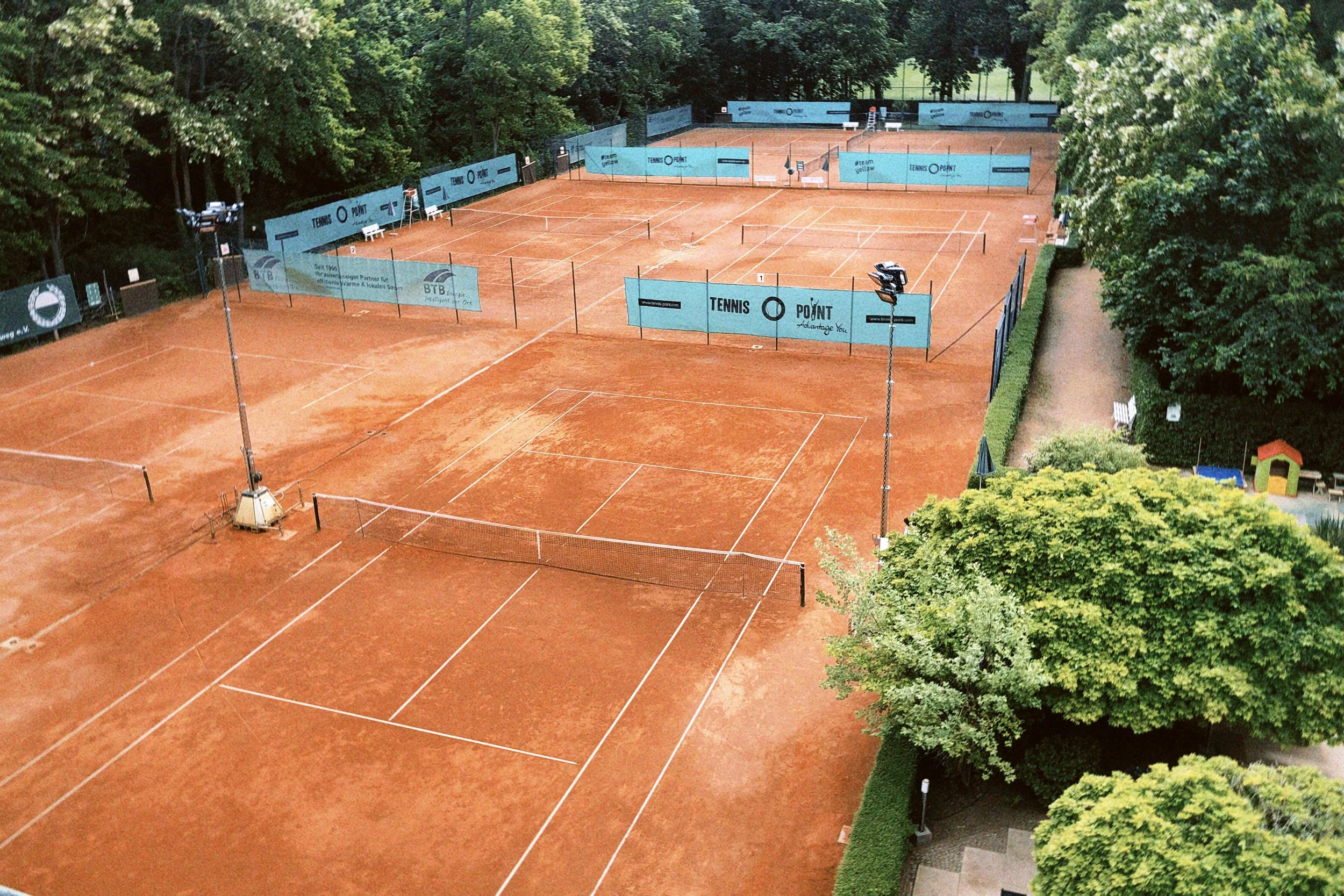 Leere Tennisplätze mit roten Sandbelägen, umgeben von grünen Bäumen und Sichtschutzwänden.