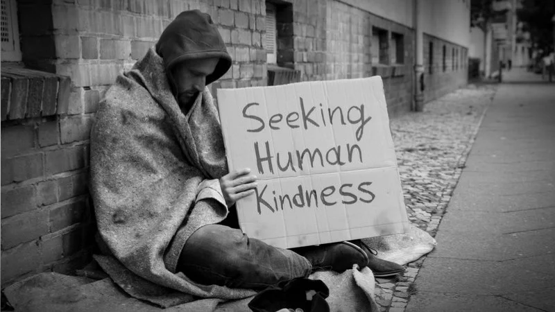 Homeless man covered with blanket holding a sign that states "seeking human kindness".