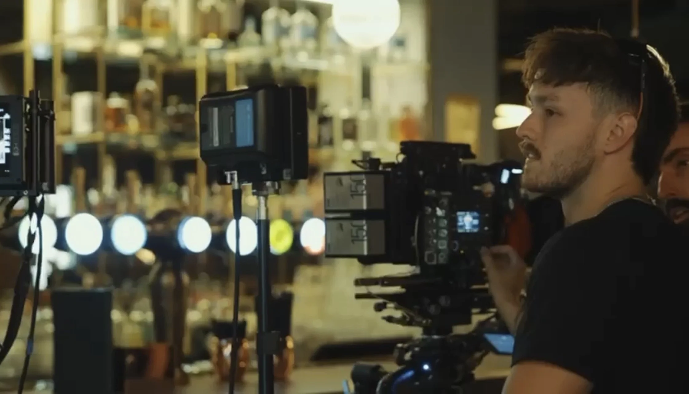 A man filming a scene with a professional camera setup in a bar or restaurant, with shelves of bottles in the background.