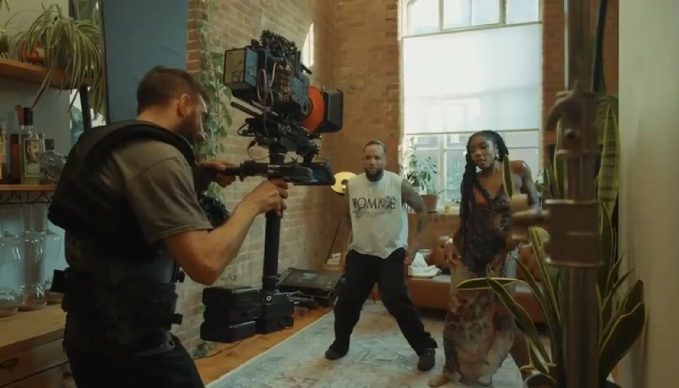 Filmmaker operating camera on set with two actors in a well-lit room with large window, brick walls, and houseplants.