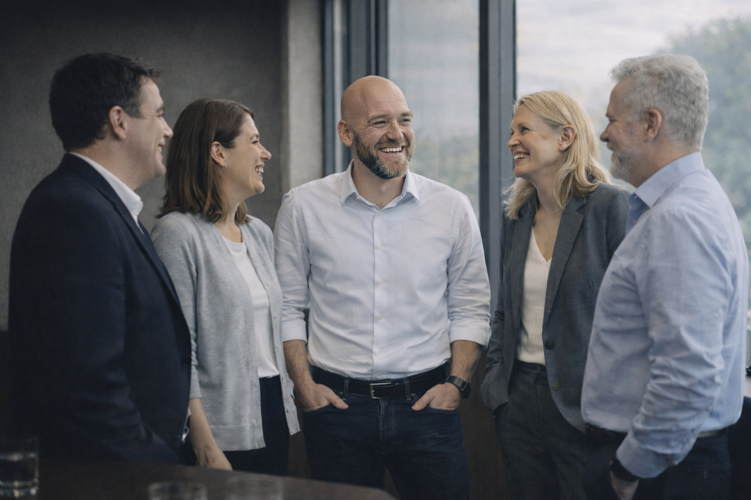 Five professionals in business attire smiling and talking in a conference room with large windows.