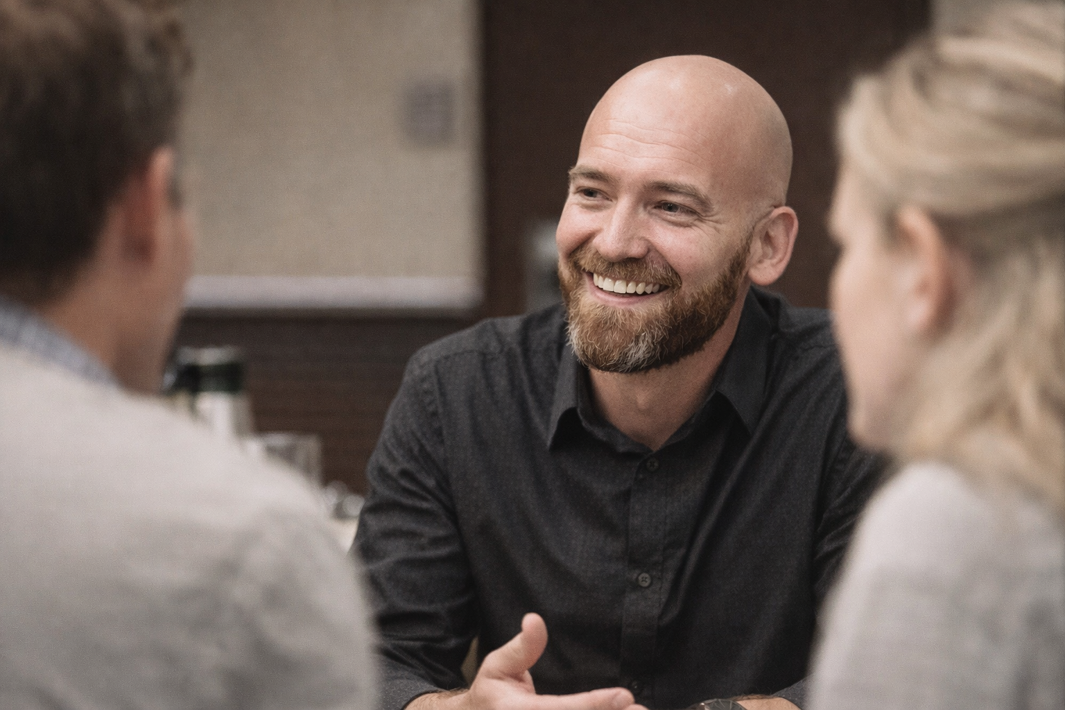 A smiling, bald man with a beard, wearing a dark shirt, talking to two people in a cozy indoor setting.