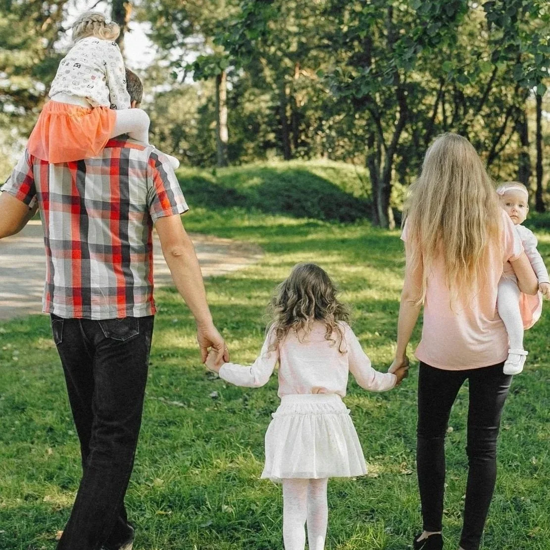 Family walking outdoors in a park, with two women, two young girls, and a man, all holding hands and enjoying a sunny day.
