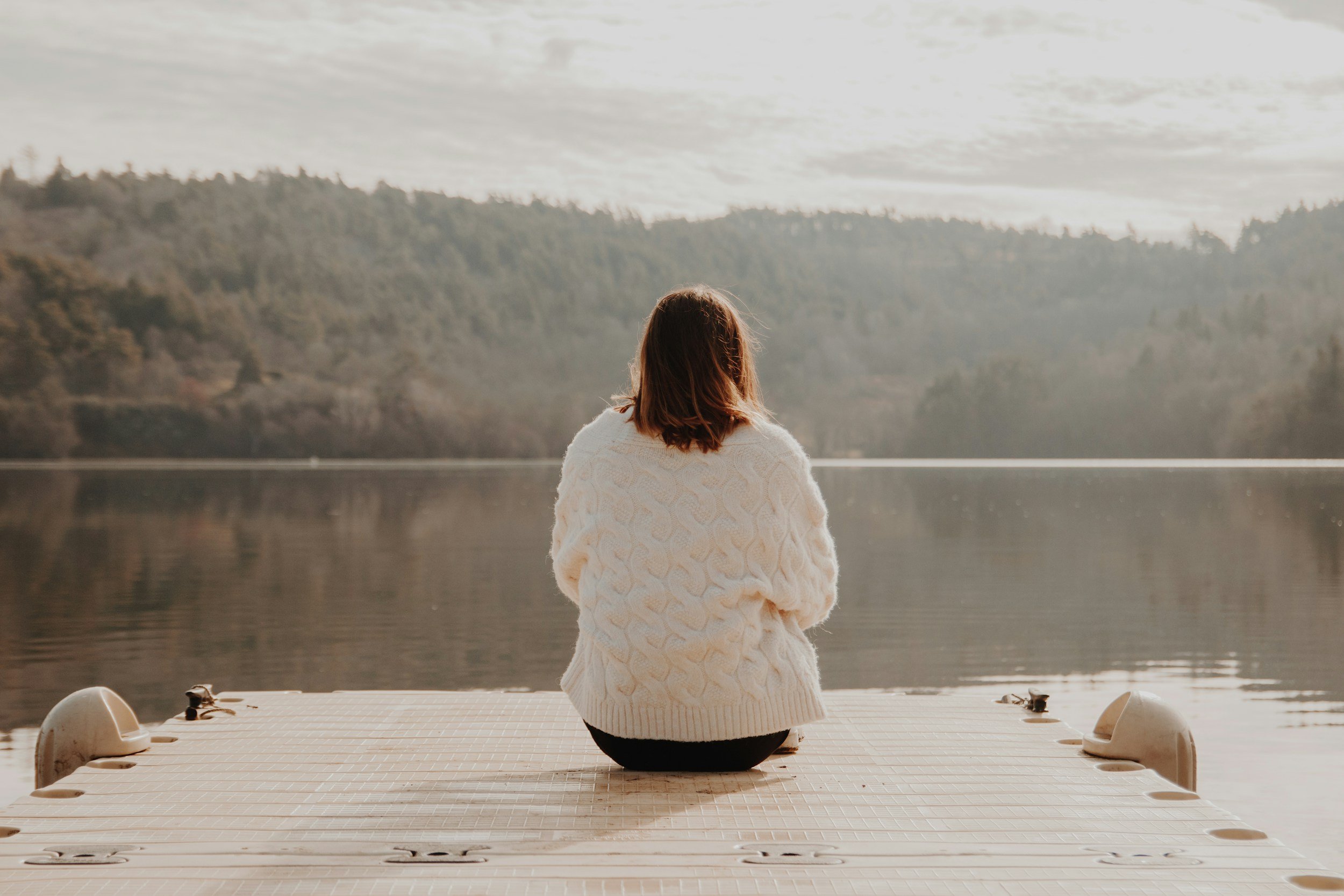 A person with shoulder-length brown hair sits on a dock, facing a calm body of water with forested hills in the background, during daytime.