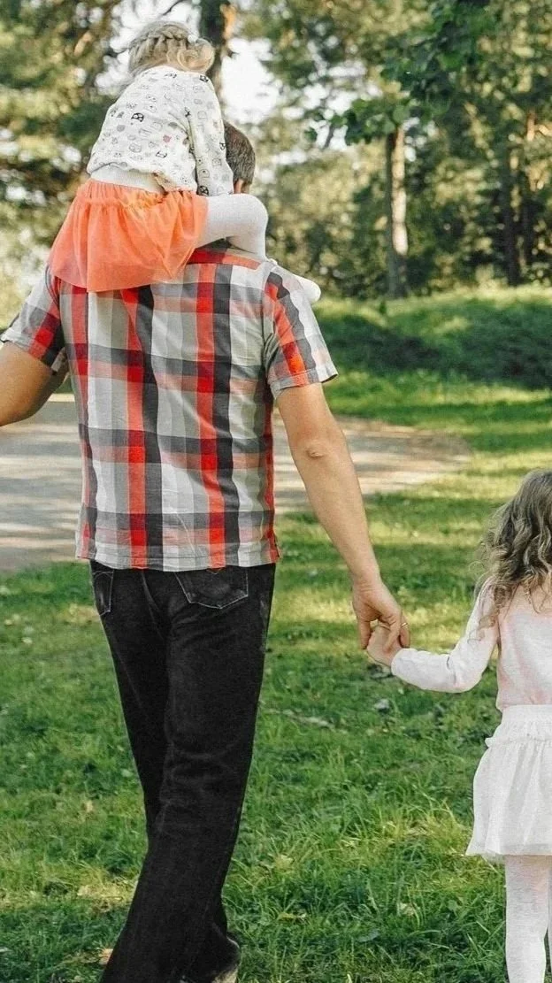 A person holding hands with a small girl while carrying a young girl on their shoulder outdoors in a park, with trees and grass in the background.