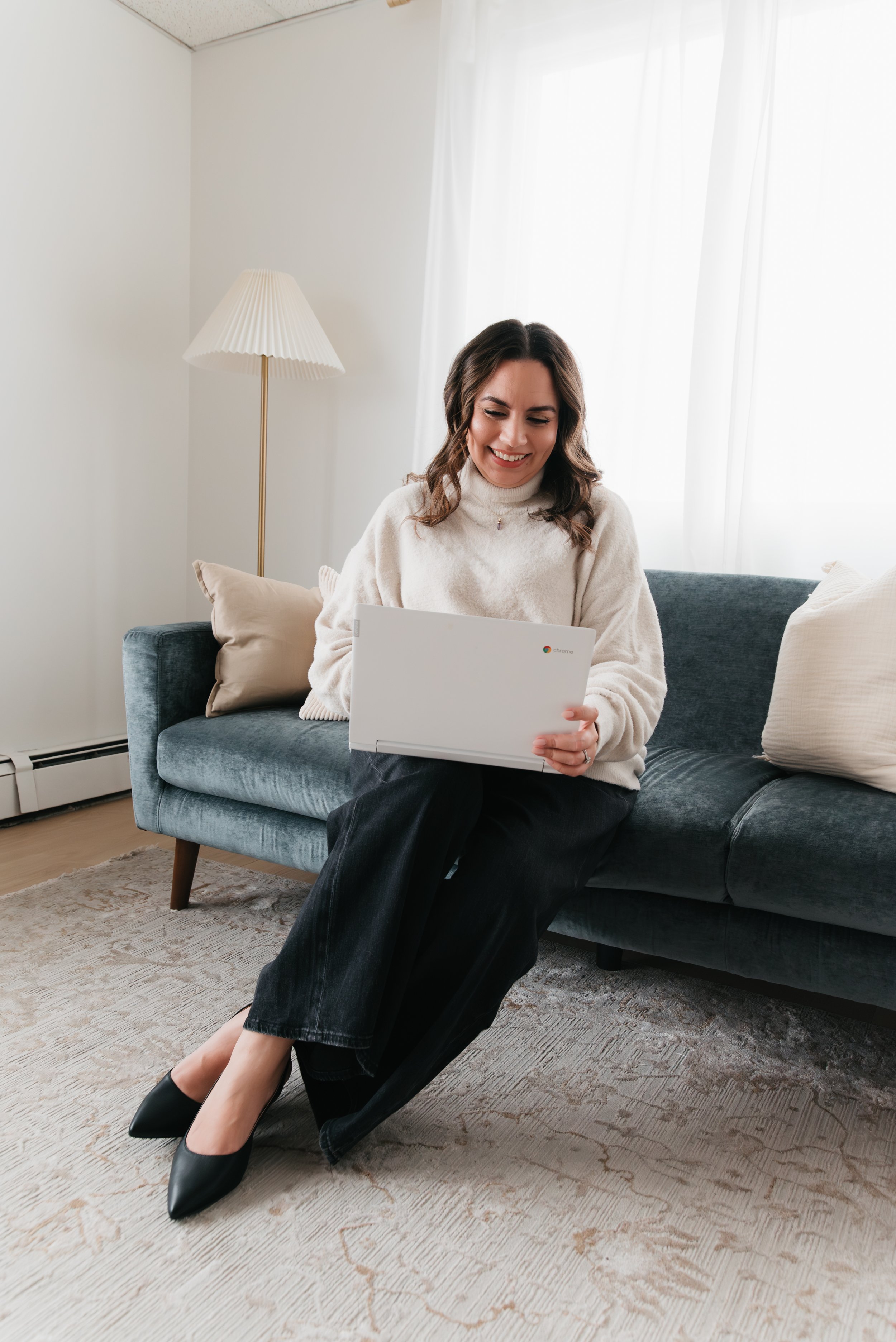 A woman sitting on a blue sofa, smiling and looking at a Chromebook. She has dark hair, is wearing a cream sweater, black pants, and black high heels. The room has white walls, sheer curtains, and a beige area rug. A floor lamp with a pleated lampshade is visible behind her.