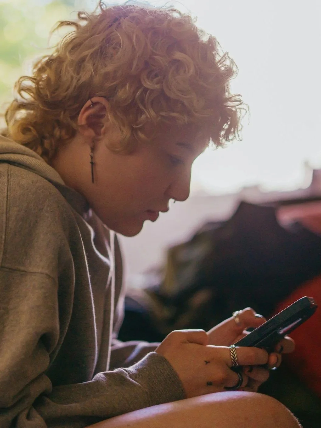 A young person with short curly blond hair looking at their smartphone, wearing multiple rings and a cross earring, in a softly lit room.