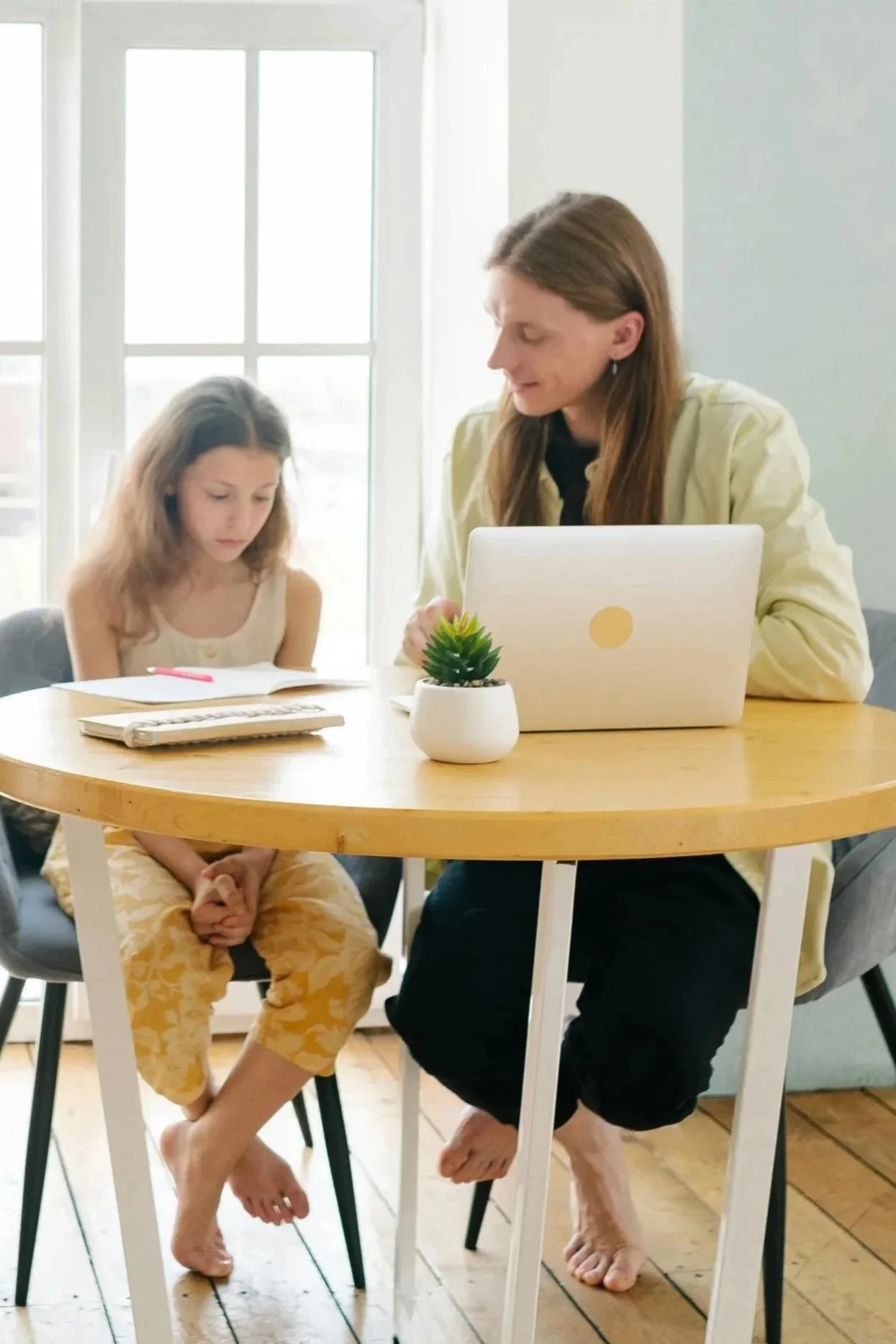A woman and a girl sitting at a round wooden table. The woman is working on a laptop, and the girl is sitting with her hands clasped in her lap, looking down. There is a small potted plant on the table. They are both barefoot, with the girl wearing yellow patterned pants and the woman wearing black pants and a light yellow jacket. Behind them, large windows let in bright natural light.