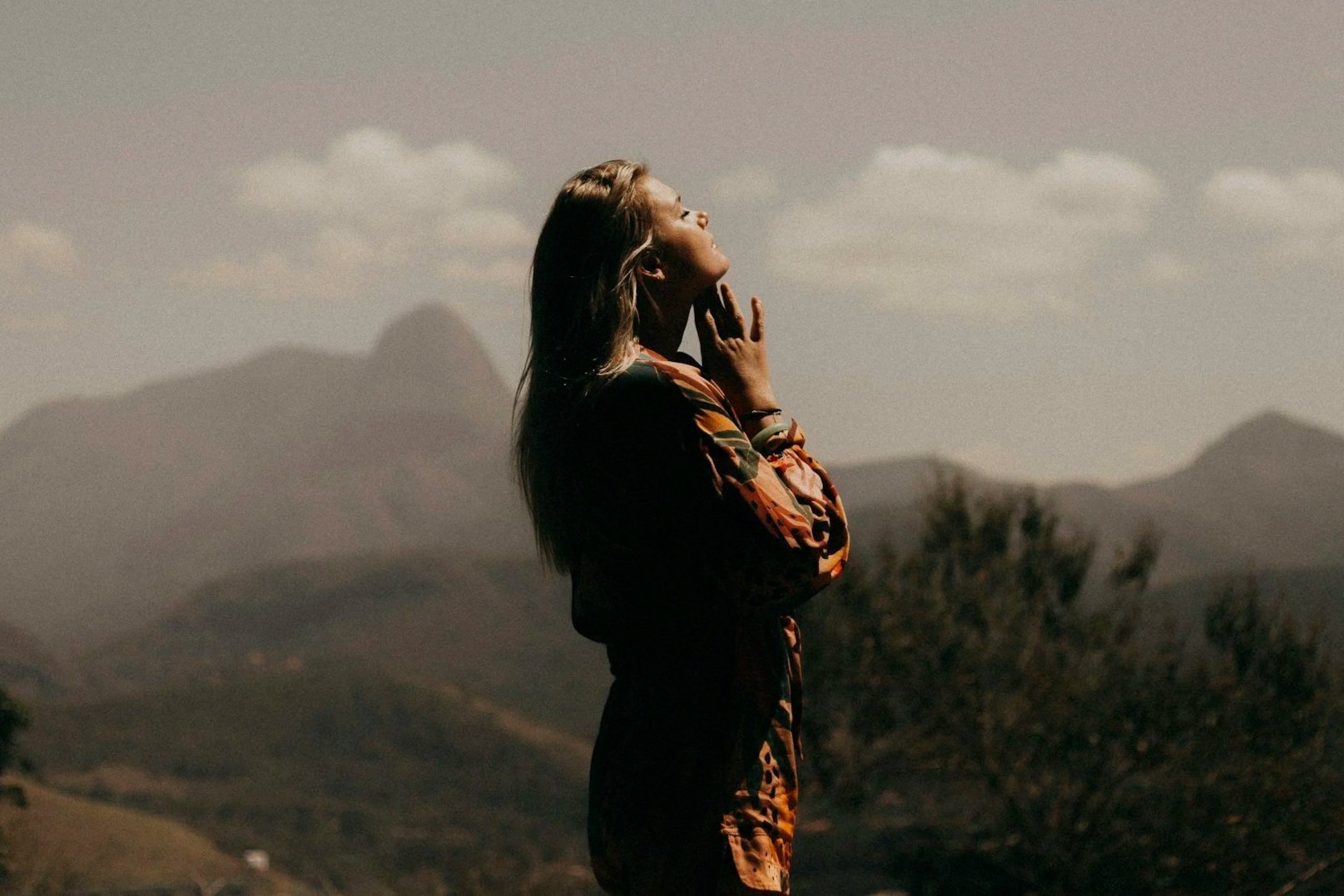 A woman with long hair standing outdoors, looking upward with her hand on her chin, in a scenic landscape with mountains and trees in the background.