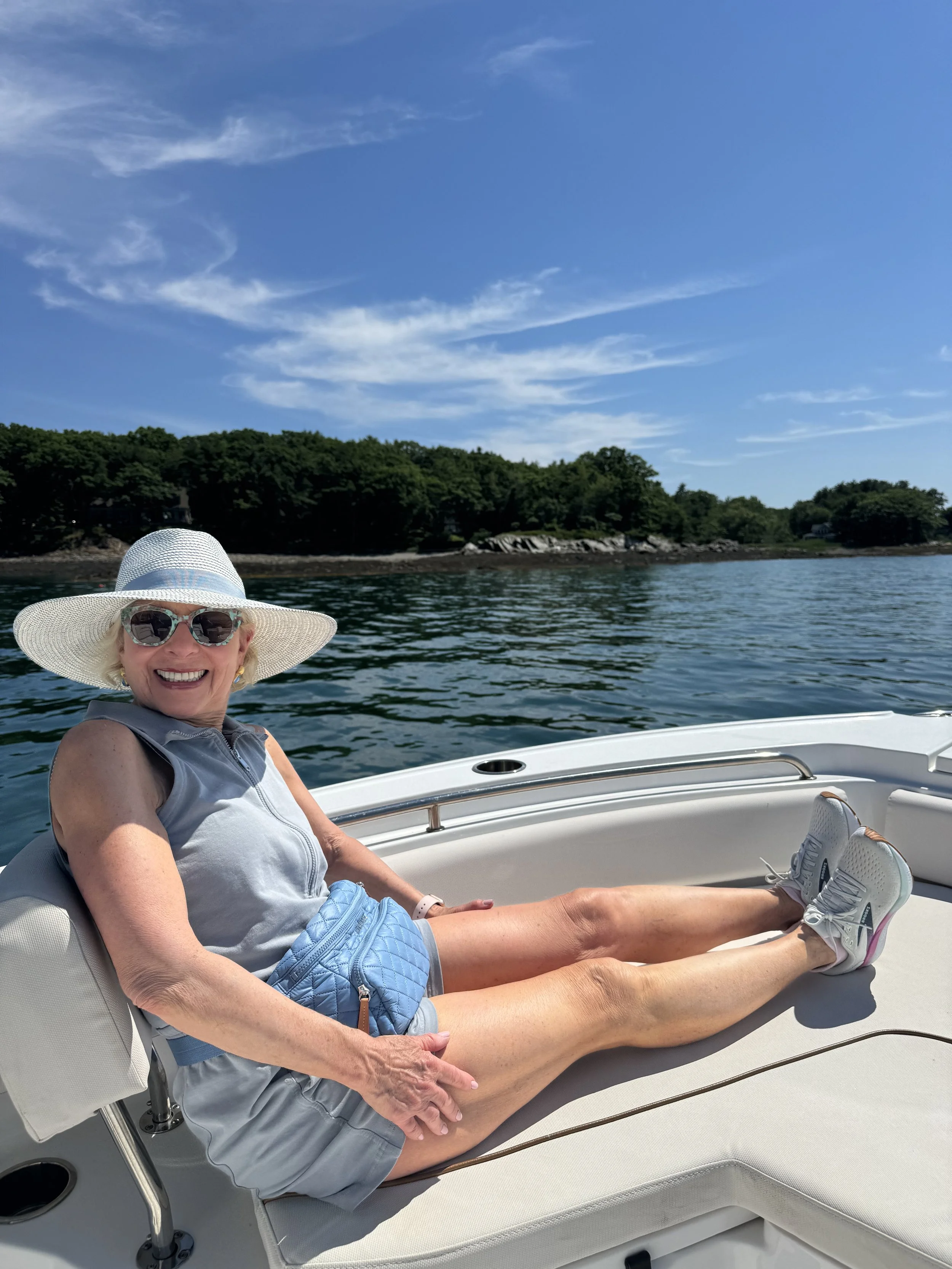 Smiling elderly woman wearing a large sun hat, sunglasses, a sleeveless gray top, and gray shorts, relaxing on a boat with clear blue sky, water, and green trees in the background.