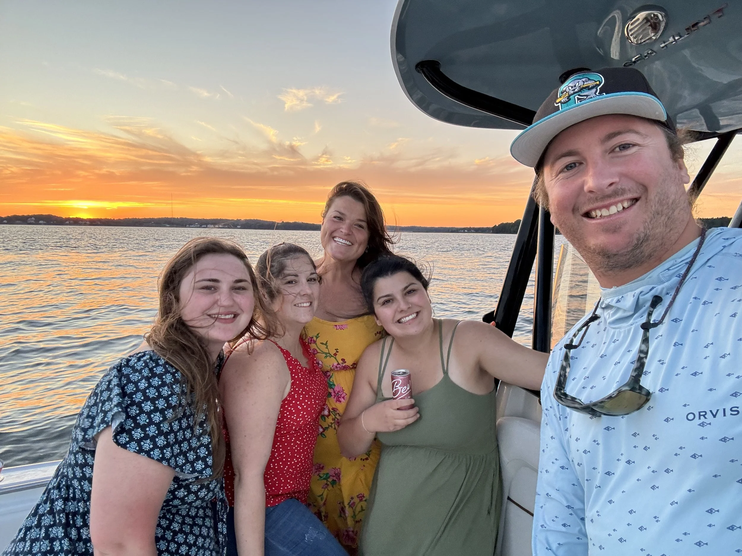 Bird Eye maine five people smiling on a boat during sunset with water and a colorful sky in the background.
