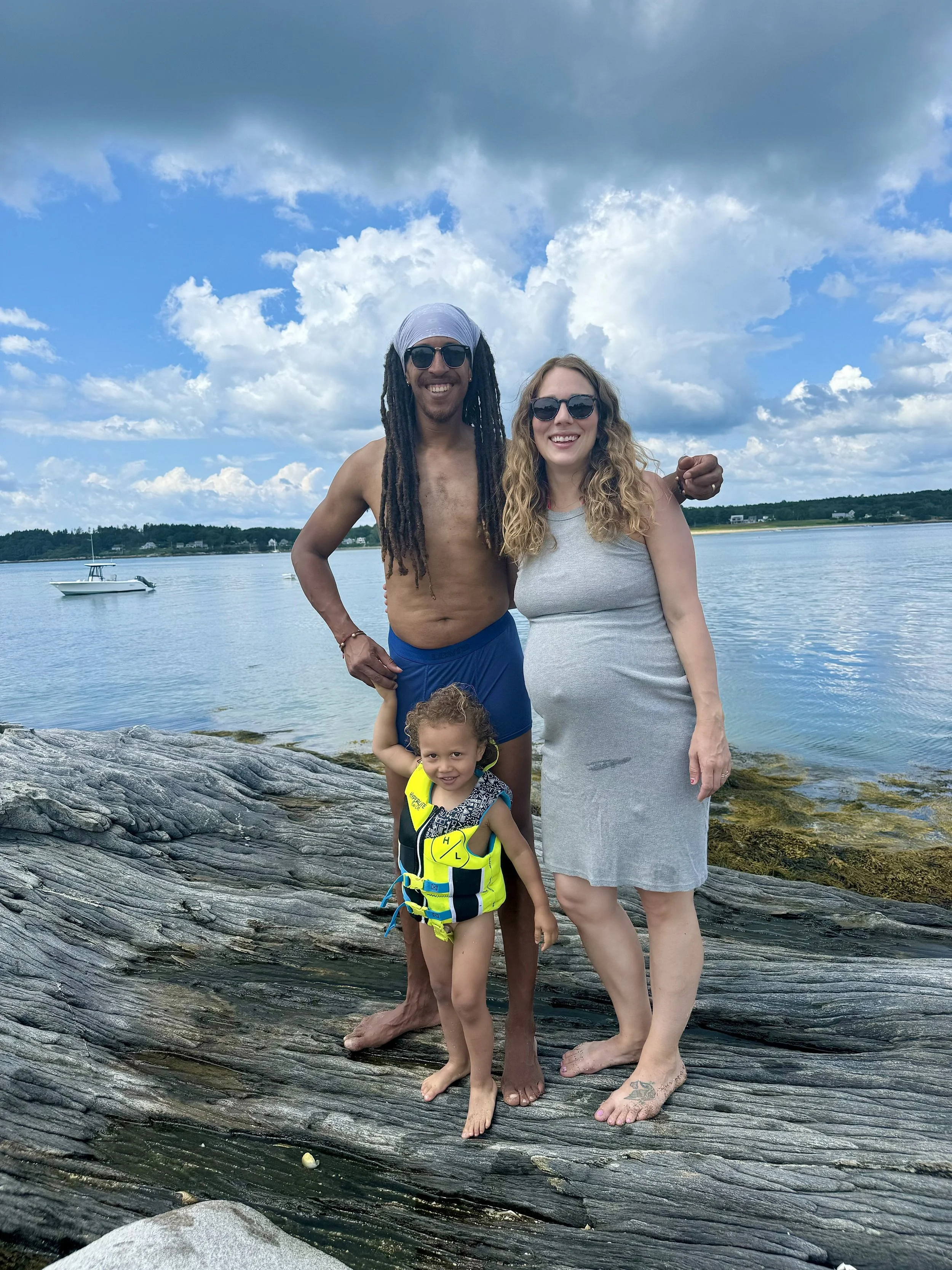 Family of three, a man, woman, and young girl, standing on rocks by a body of water with a boat in the background, under a cloudy sky.