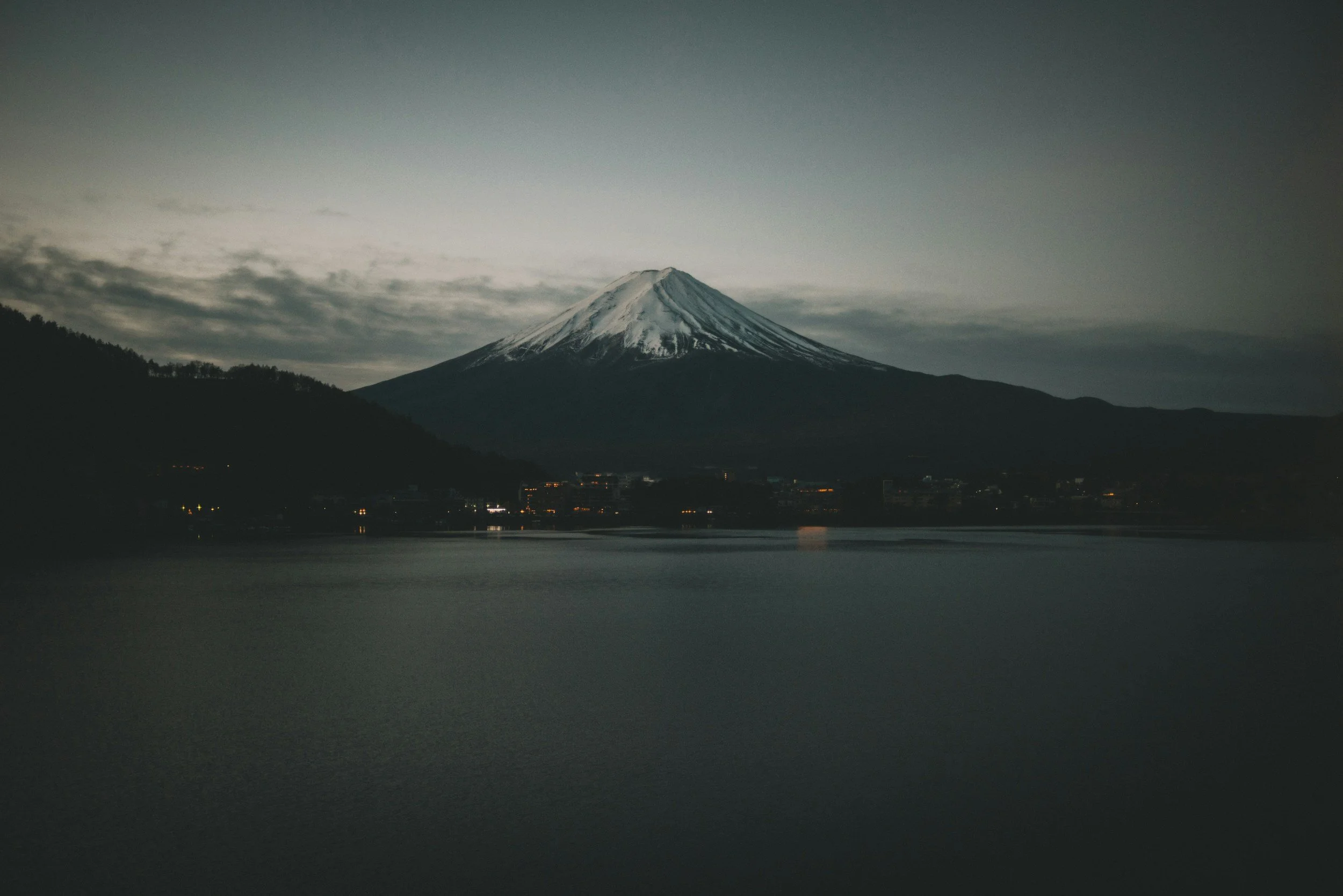 Mount Fuji at dusk with snow-capped peak, dark sky, and reflective lake in foreground.