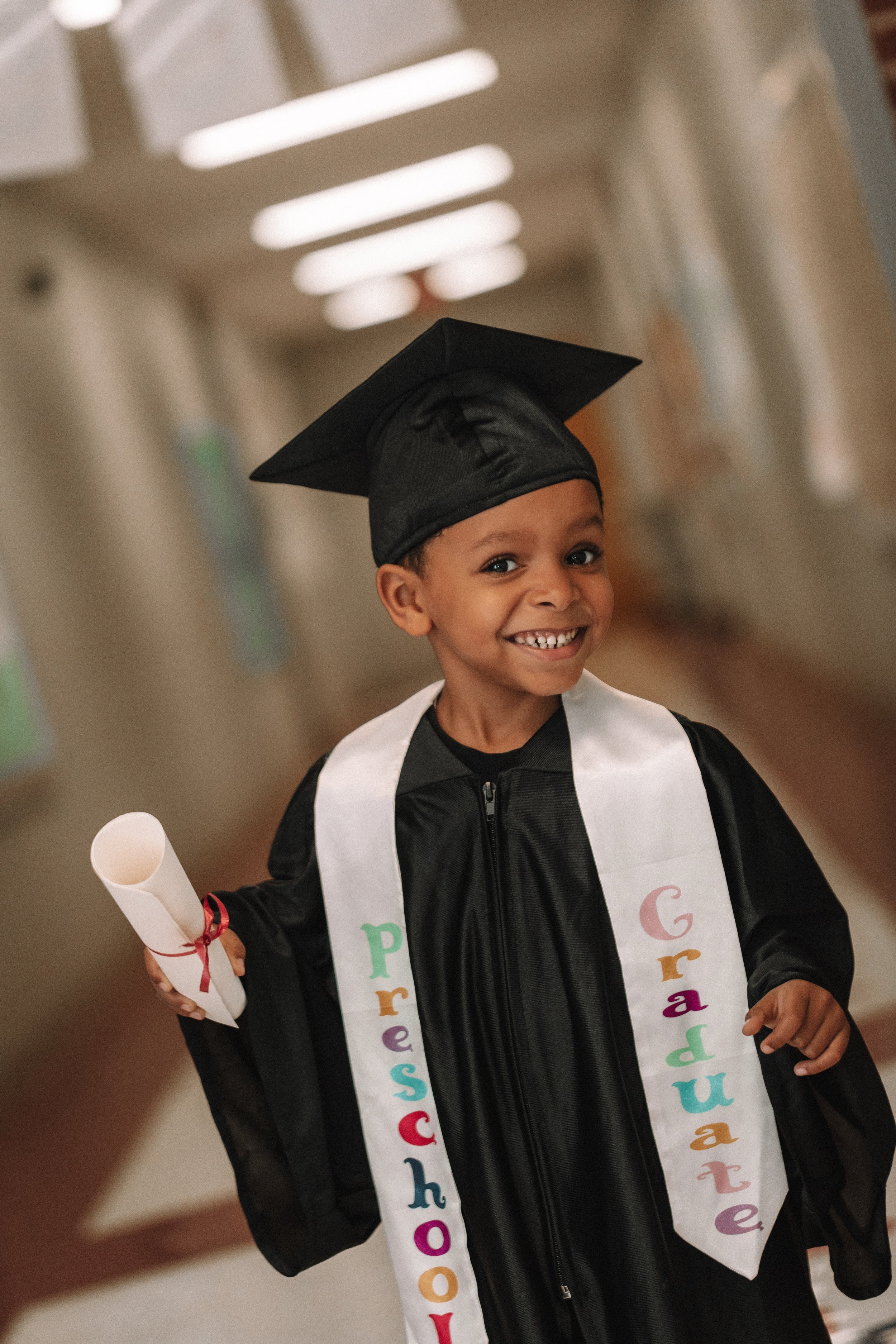 Child wearing a graduation cap and gown, holding a diploma, with a sash labeled "Preschool Graduate" in a hallway.
