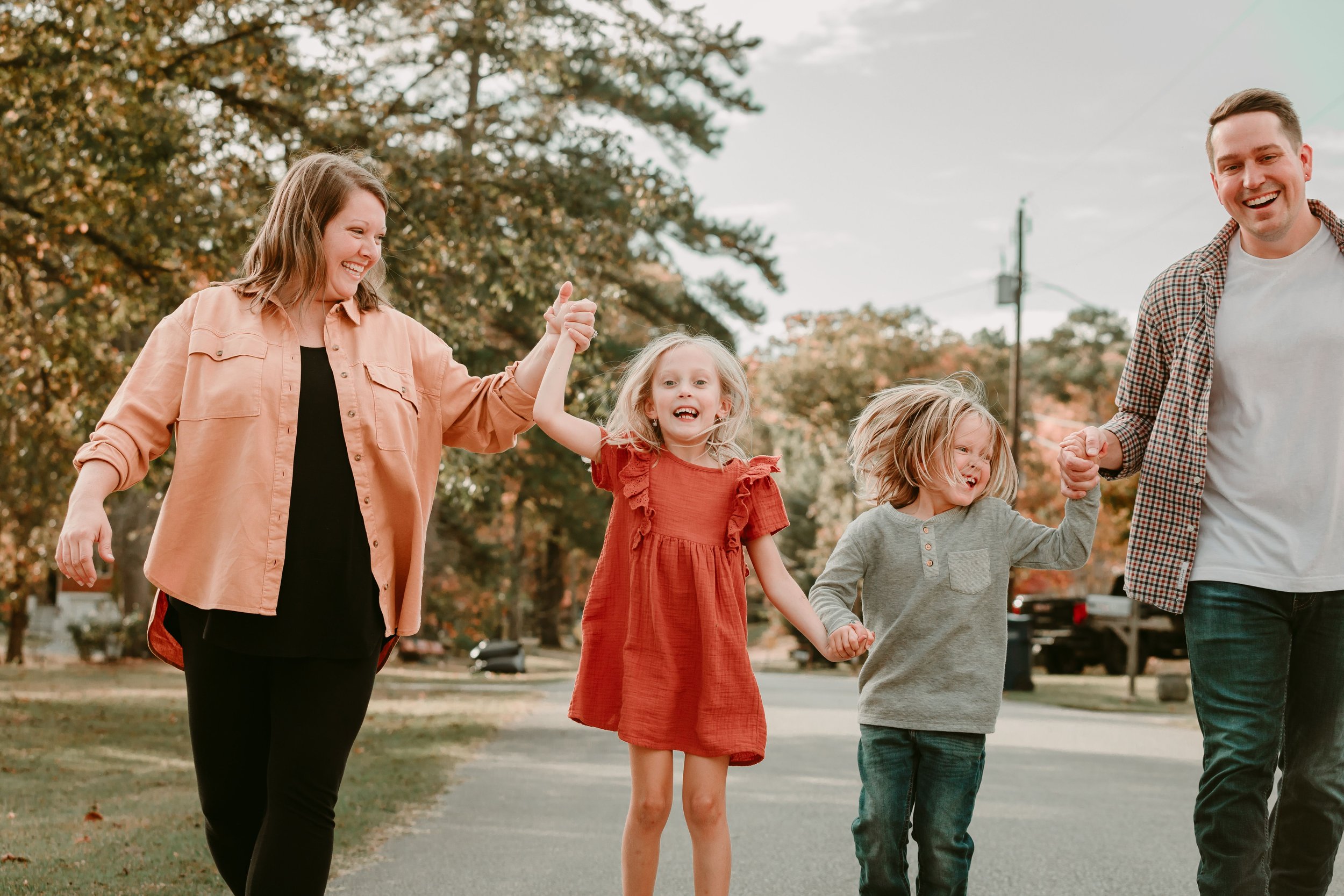 Family of four walking and holding hands outdoors, smiling in a scenic park setting with trees in the background.