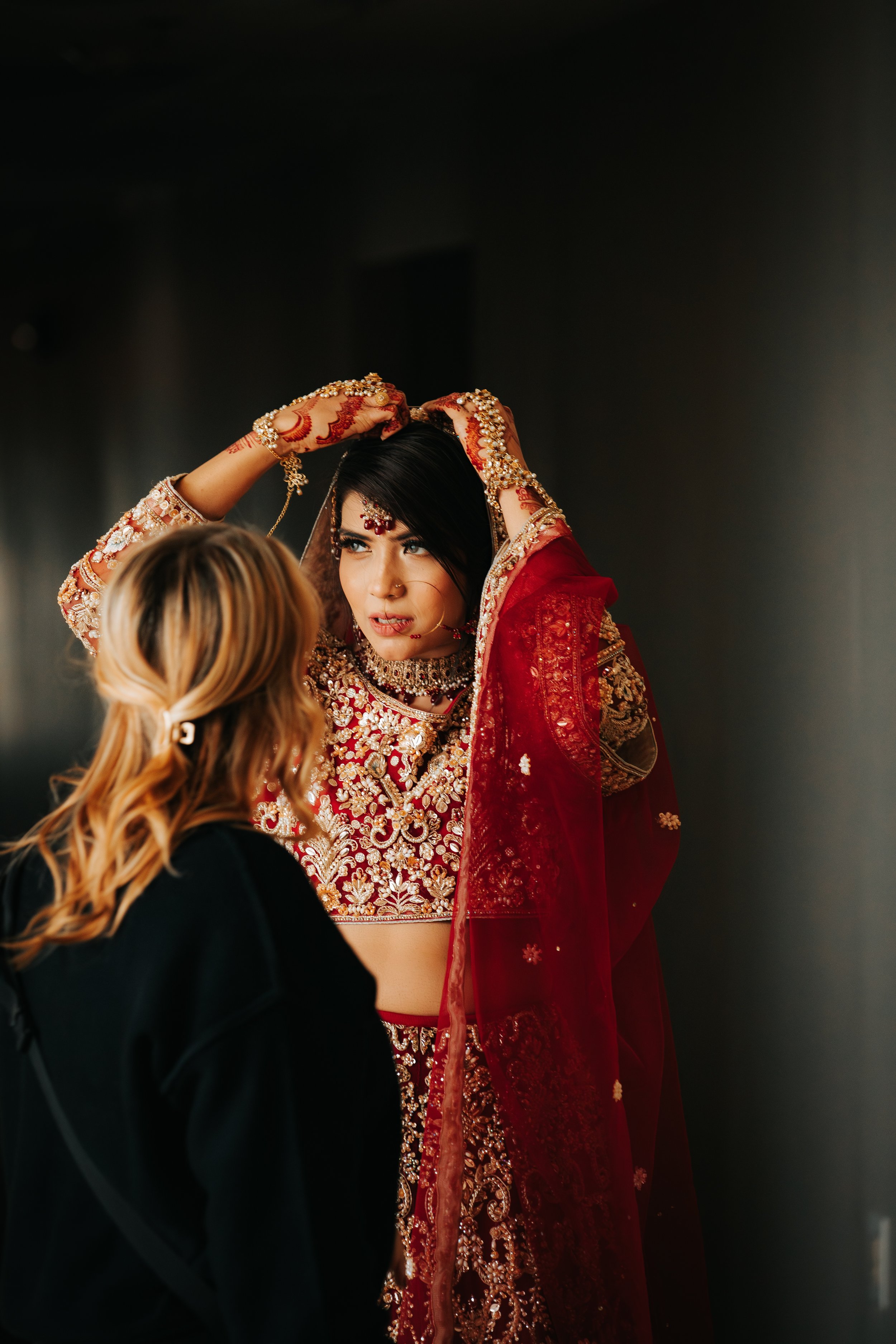 A woman in an elaborately embroidered red bridal outfit, adjusting her jewelry with the help of another person.