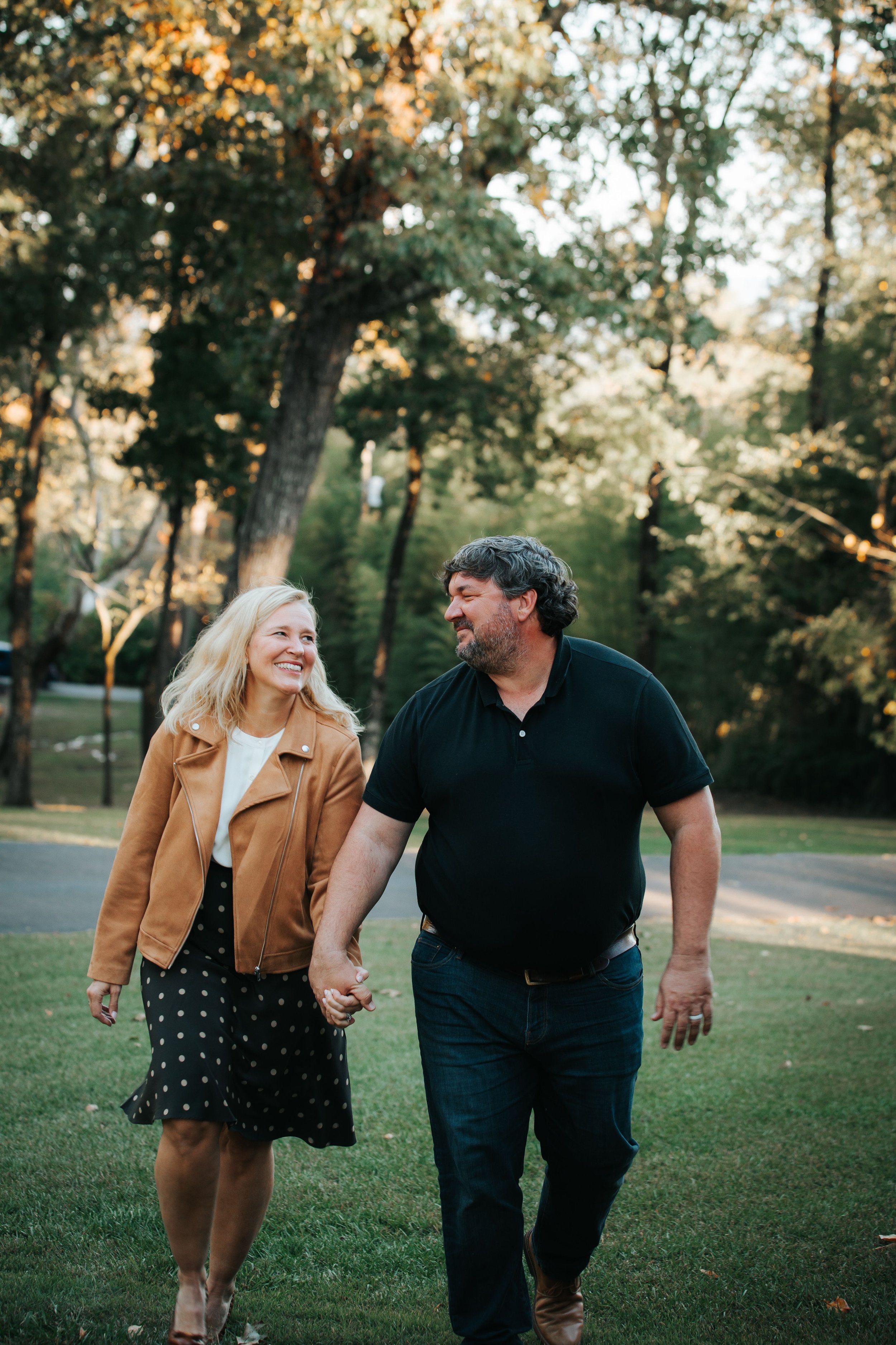 A couple walking hand in hand on a grassy path with trees in the background, smiling and looking at each other, during daytime.