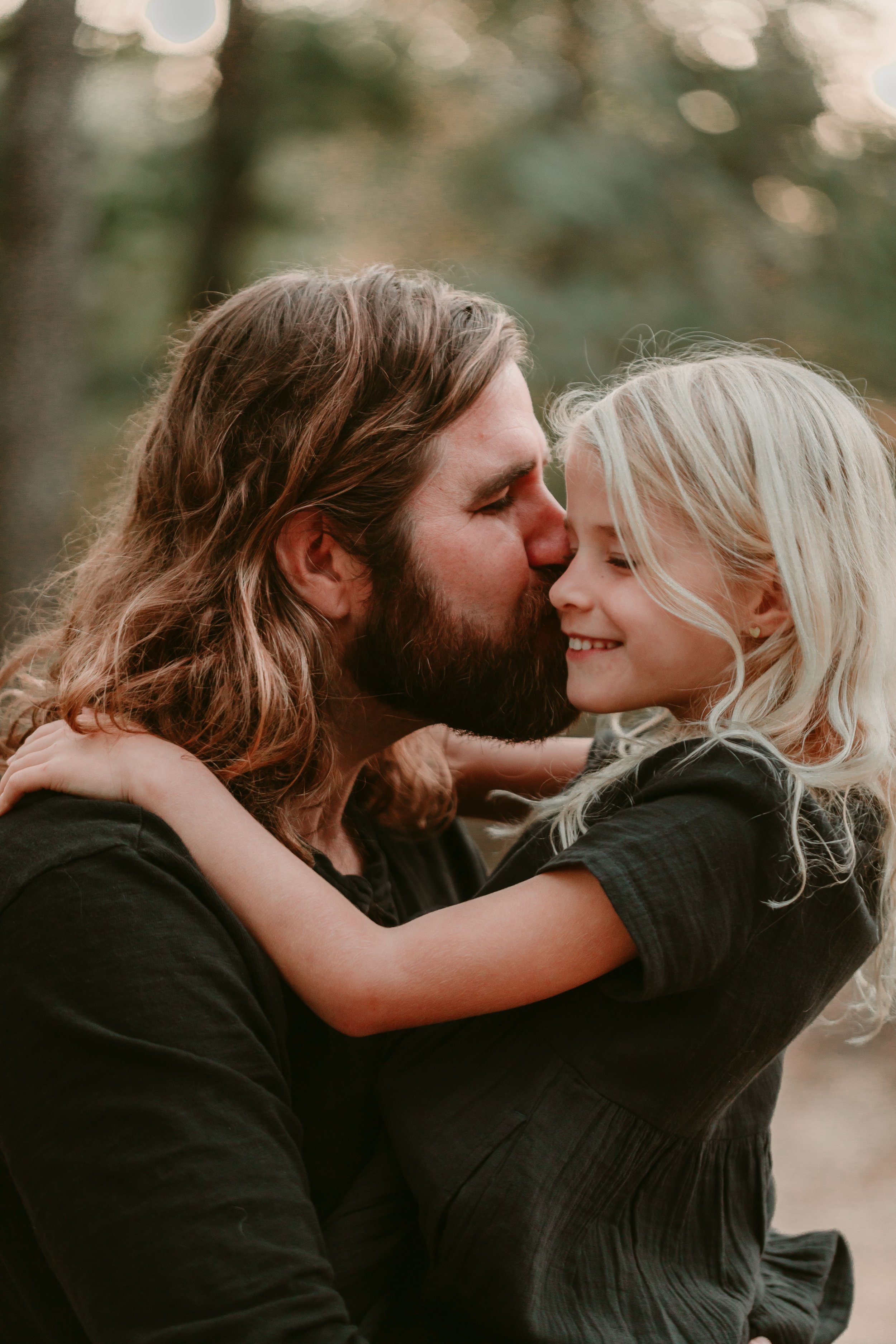 A man with a beard kissing a smiling young girl on the nose in a tender moment outdoors, both wearing dark clothing.