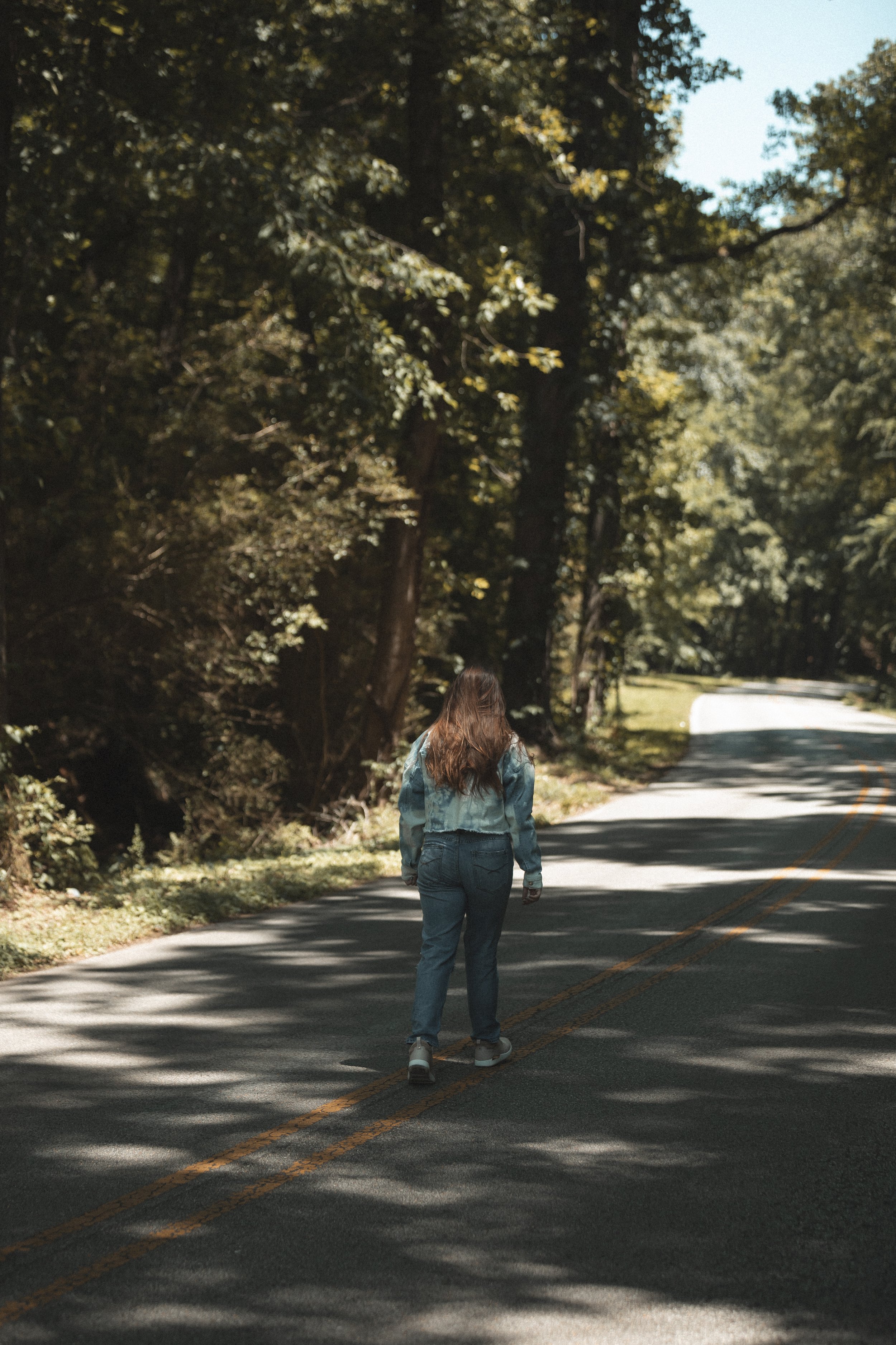 Person walking on a tree-lined road, wearing denim clothing, surrounded by lush greenery.