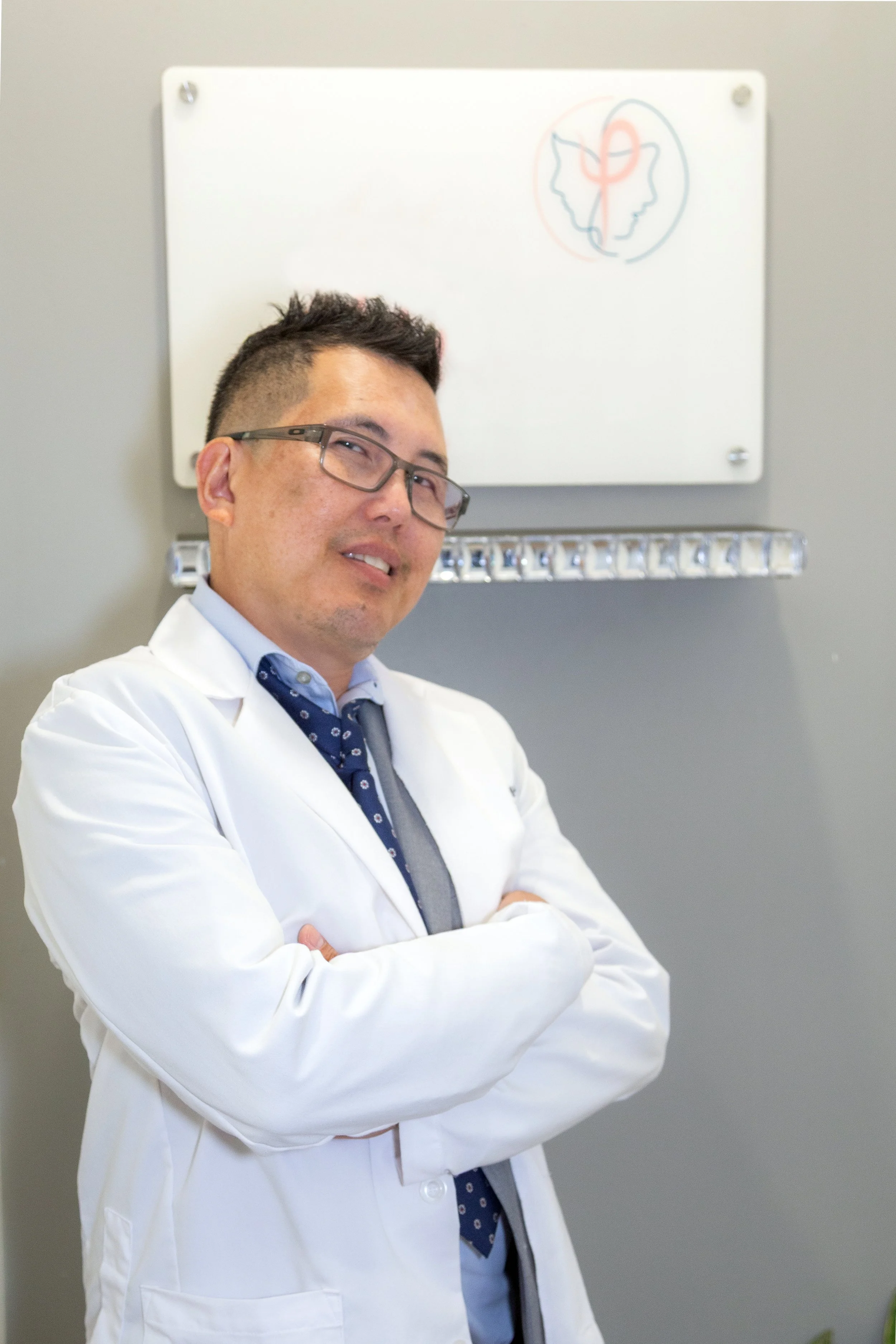 A male doctor wearing glasses, a white coat, a blue shirt, and a grey tie standing with arms crossed in front of a whiteboard with a diagram of two faces and a circle in between.
