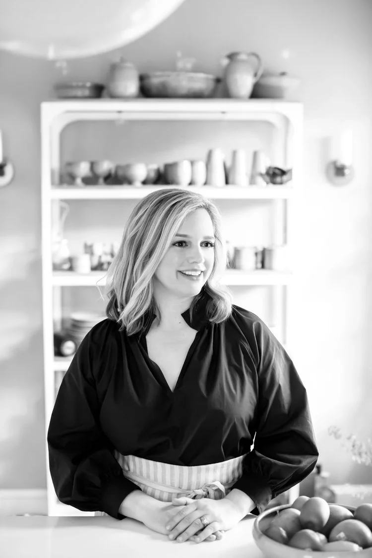 Black and white photo of a woman with blonde hair wearing a dark blouse and striped apron, standing in a kitchen with a shelf of pottery in the background.