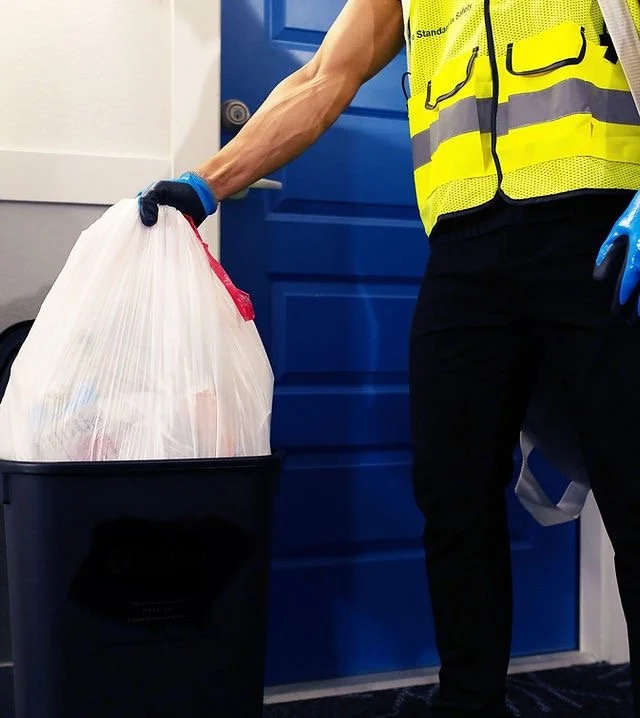 Person wearing a yellow safety vest and black pants disposing of a white trash bag into a blue recycling bin near a blue door.