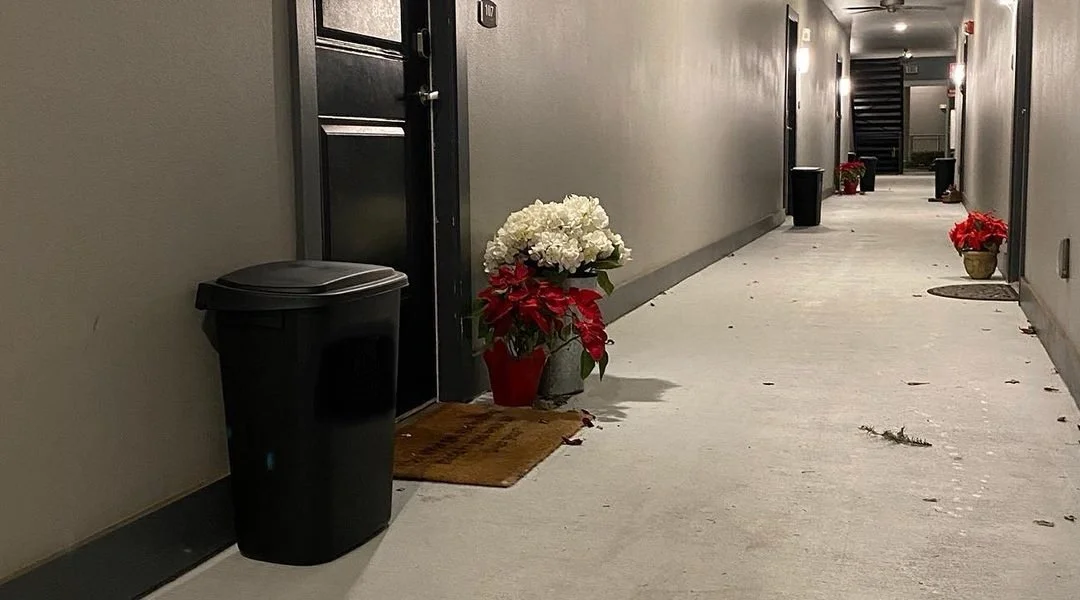 Hallway decorated with poinsettia plants and a doormat outside a door, with a trash can nearby.