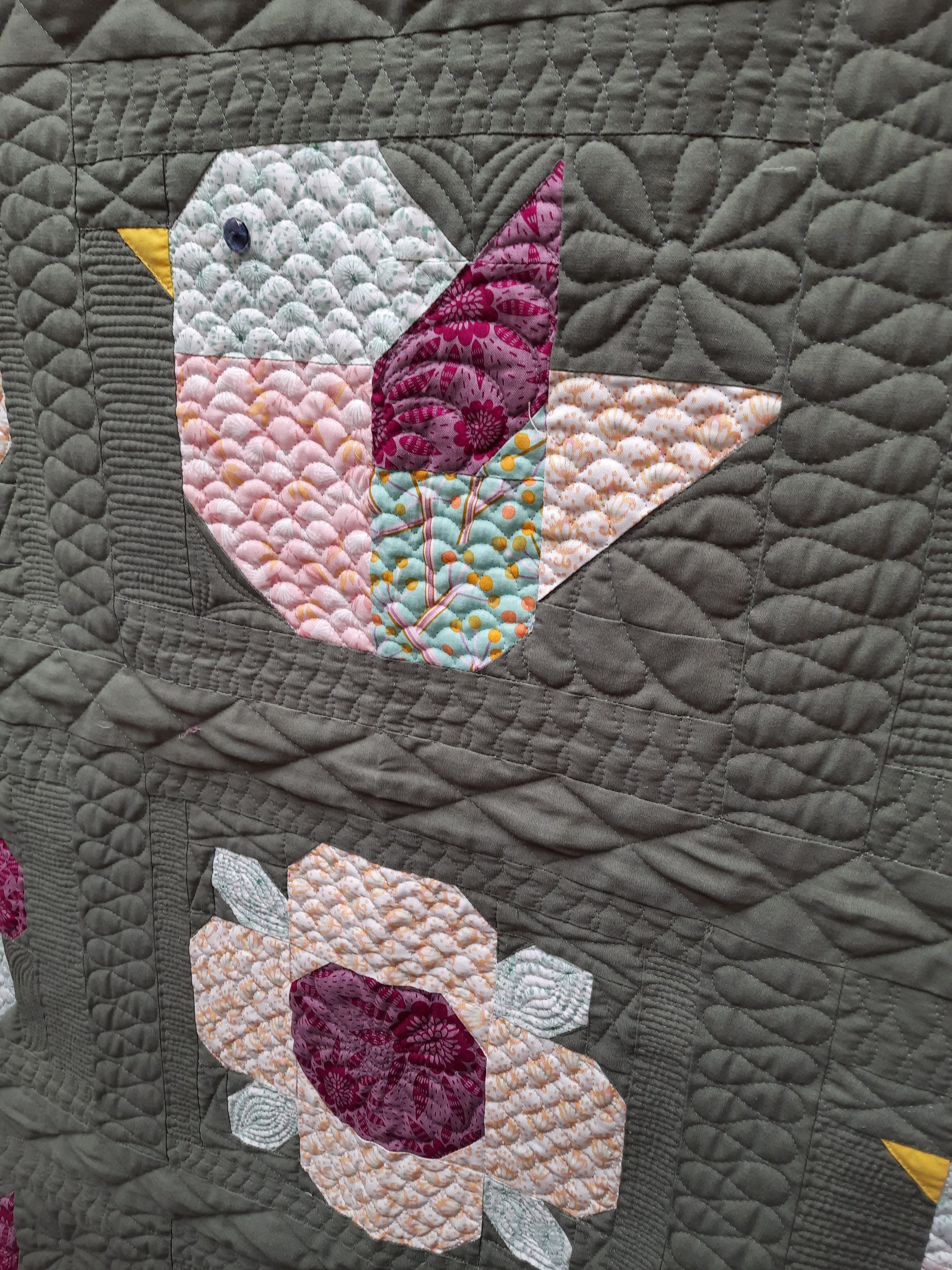 Un cojín o colcha de retazos con diseño de un pájaro y flores en tonos pastel y oscuros, sobre un fondo acolchado. Los patrones textiles son intrincados y coloridos.