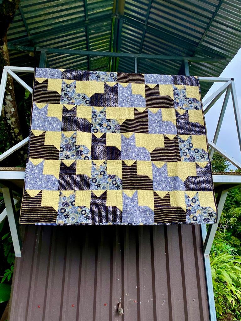 Colcha de parches con diseño de gatos colgada al aire libre, compuesta de telas en tonos amarillos, azules y marrones, con patrones de rayas y círculos.