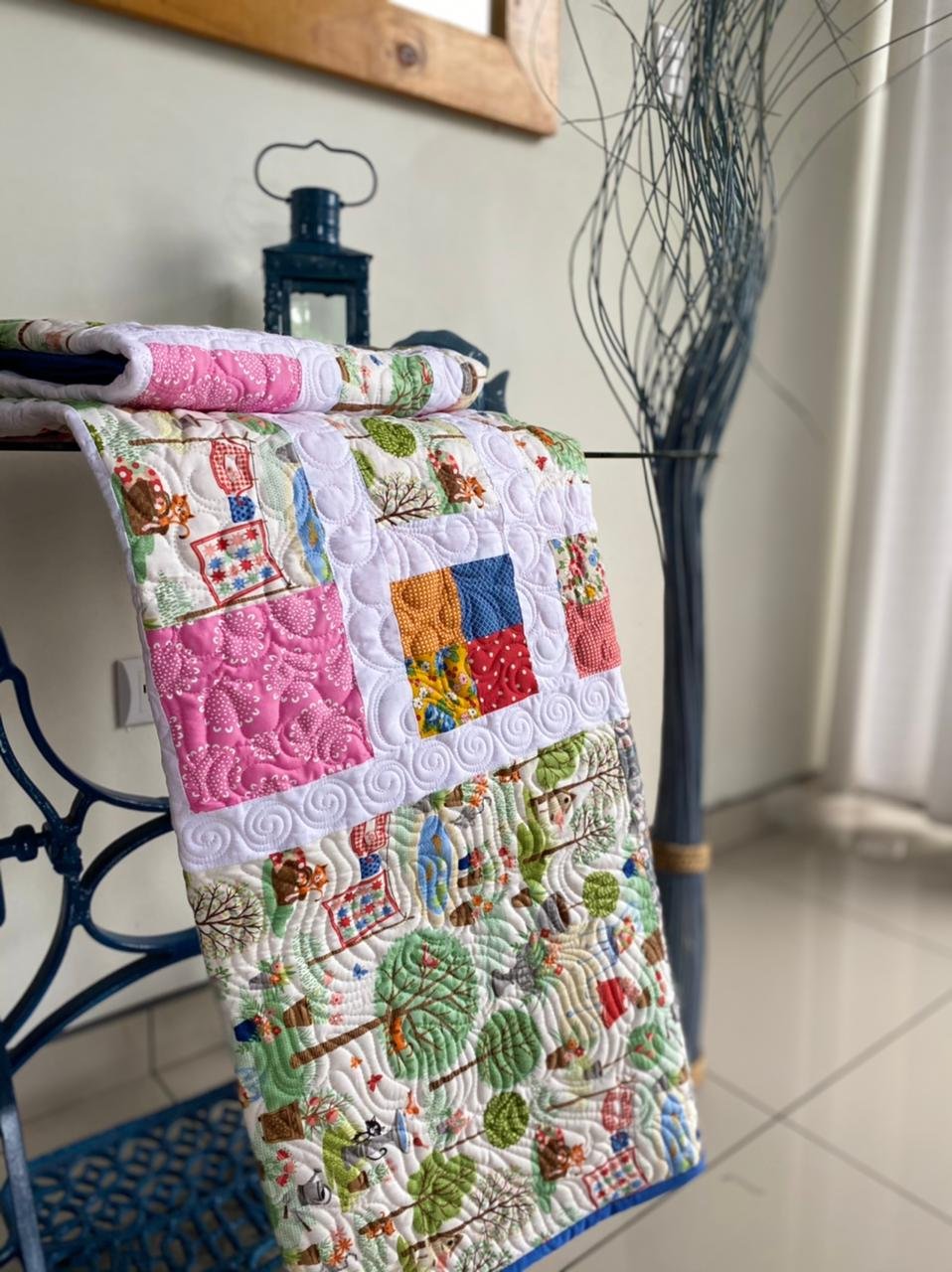 Colcha de retazos colorida sobre una silla, con diseños de árboles y animales, en un ambiente hogareño decorativo con lámpara y jarrón.