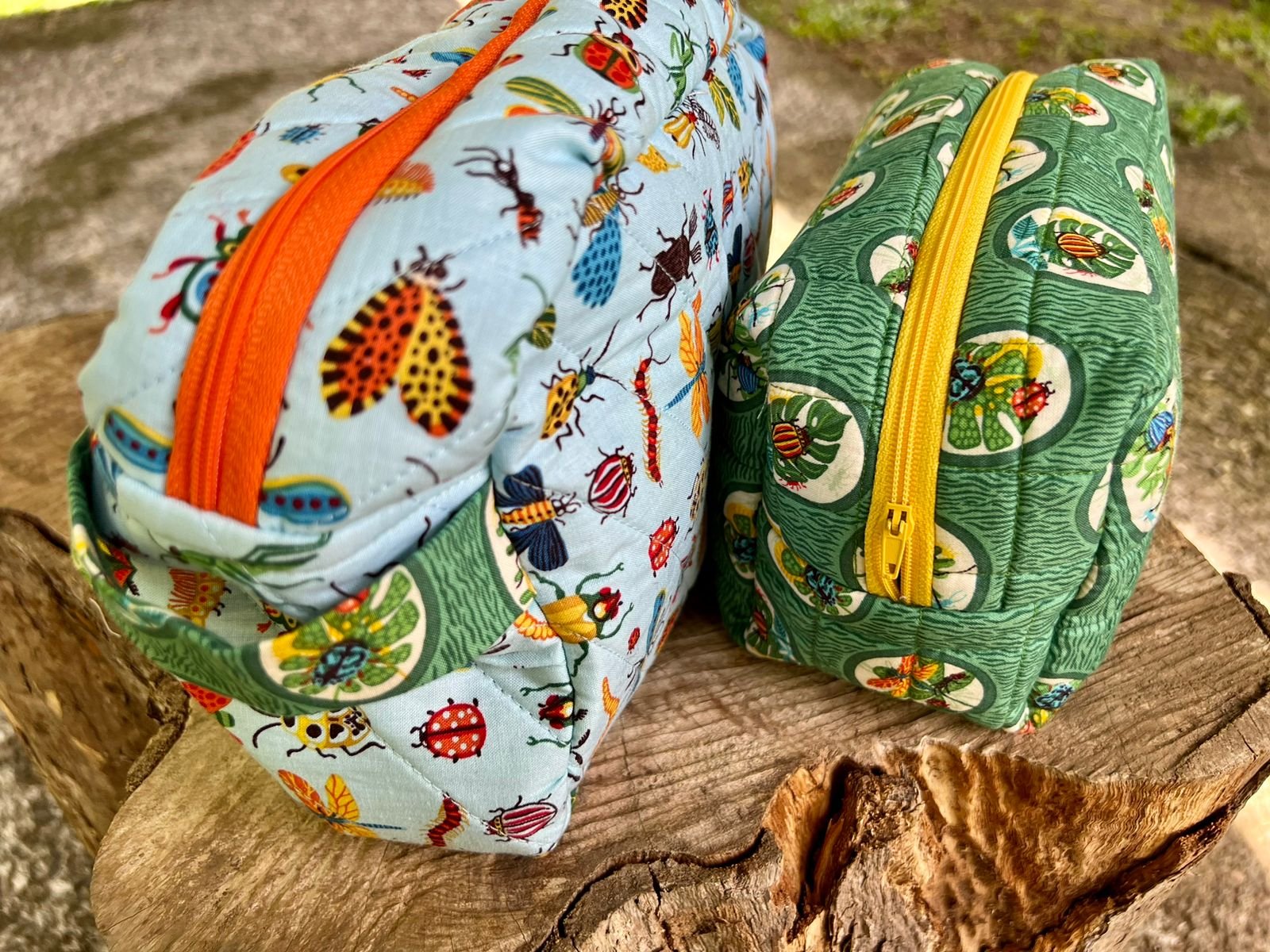 Dos estuches con diseño de insectos, uno azul claro con cremallera naranja y otro verde con cremallera amarilla, sobre un tronco.