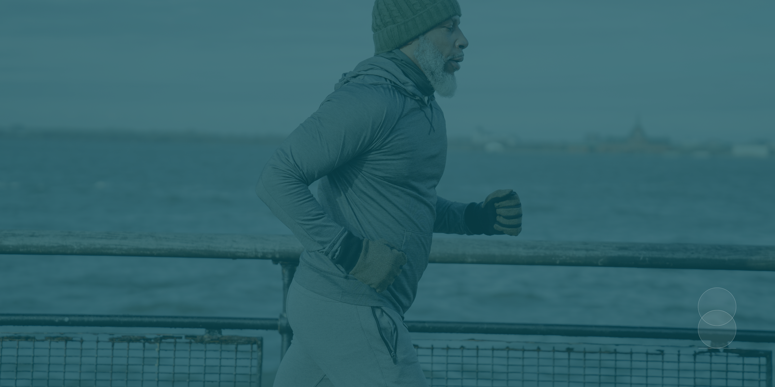 An elderly man jogging outdoors near a body of water, wearing a green knit hat, gray jacket, and gloves.