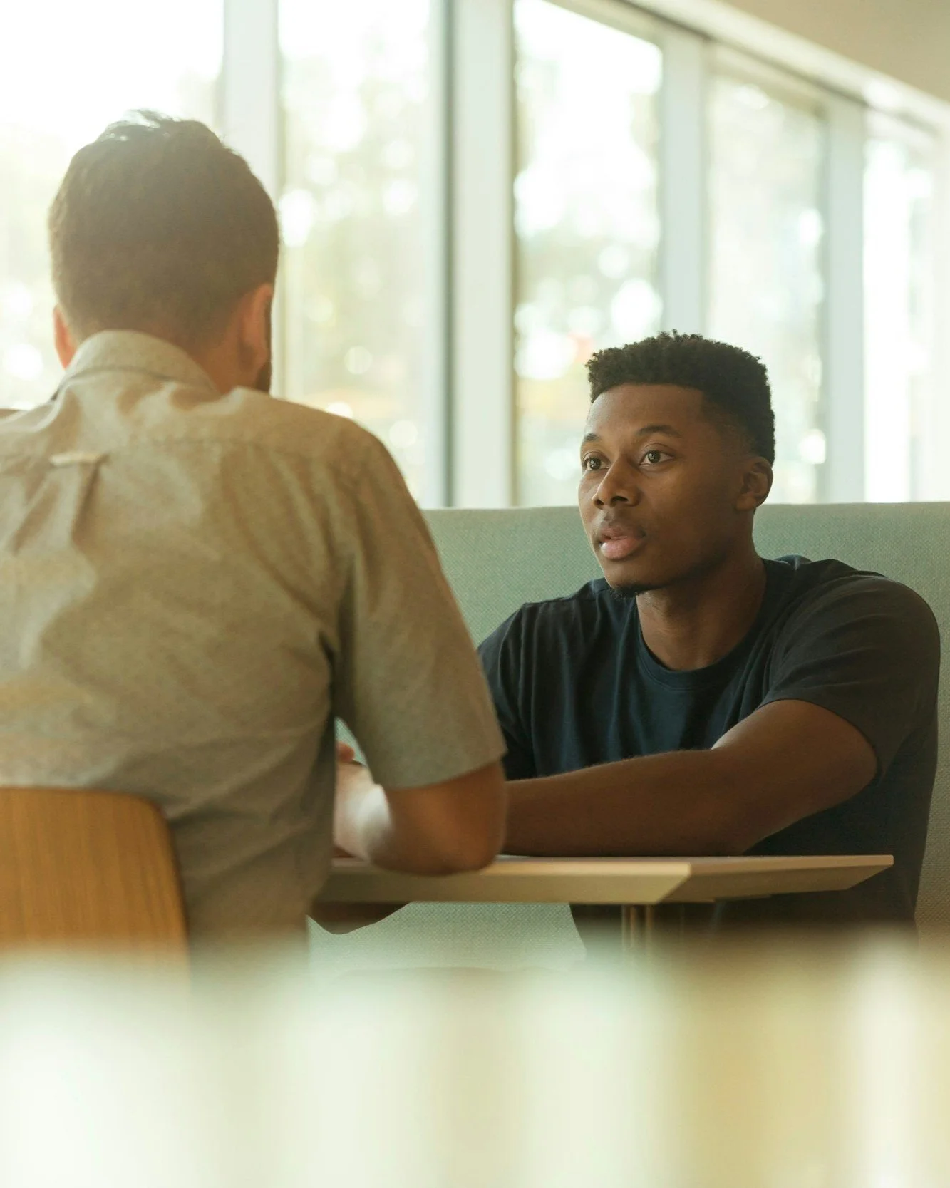Two men having a conversation at a cafe or restaurant, sitting across a table from each other with large windows in the background letting in sunlight.